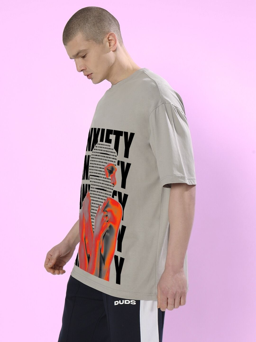 Man wearing ANXIETY OVER-SIZED T-SHIRT GREY with graphic design on front, standing against a pink background.