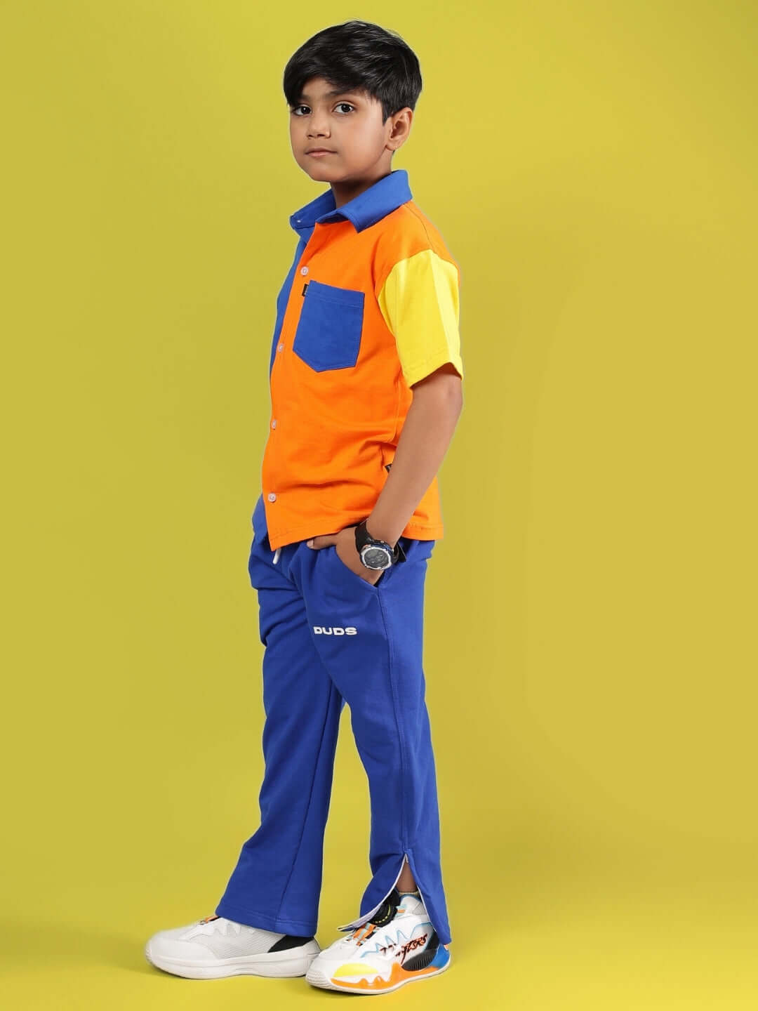 Child wearing a colorful APEX shirt and blue pants by DUDS, showcasing a vibrant multi-blue design, against a bright yellow background.