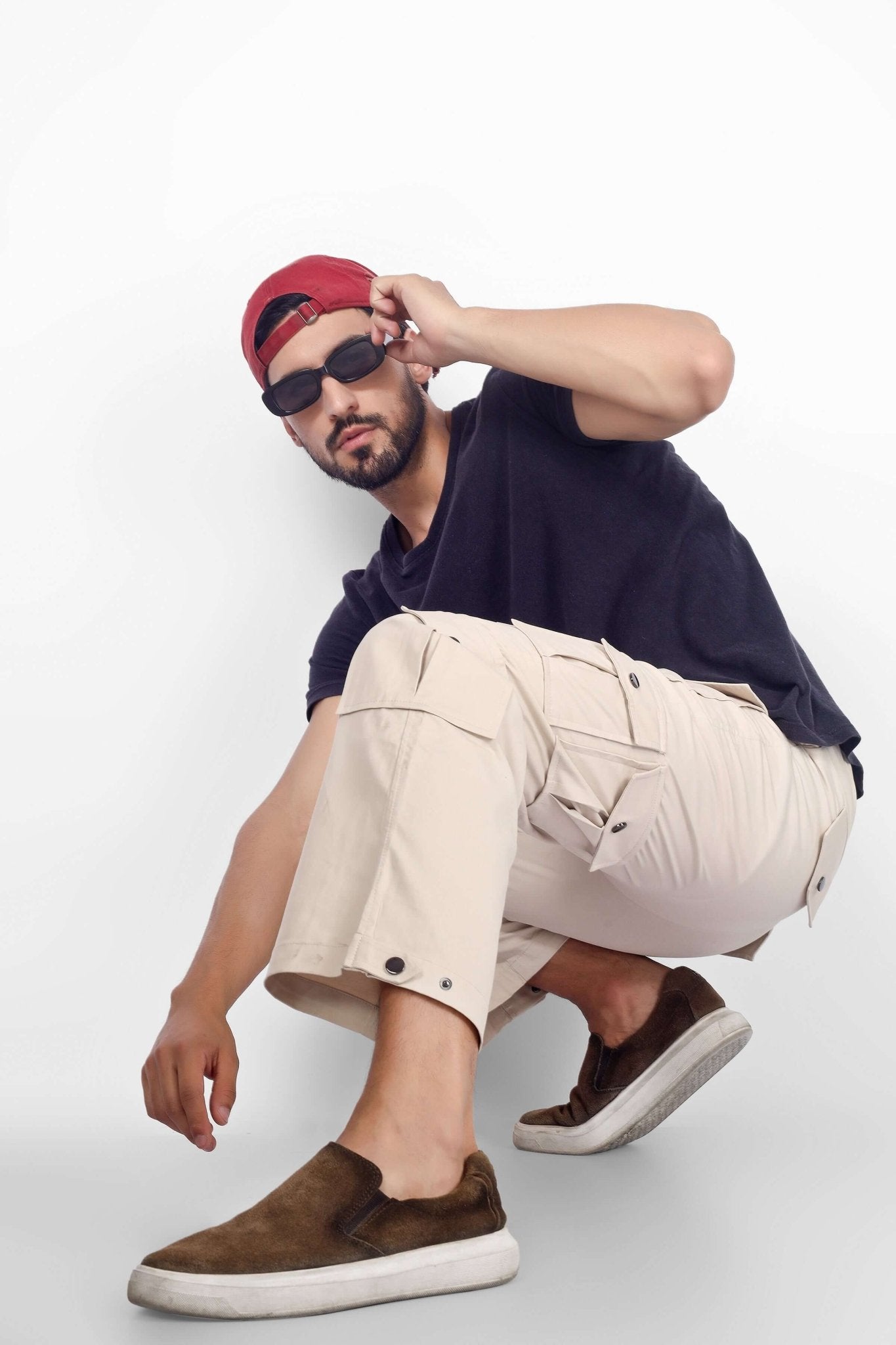 Man wearing ARCADIA WHITE 14 POCKET CARGO PANTS, squatting and posing in a casual urban style with sunglasses and red cap.