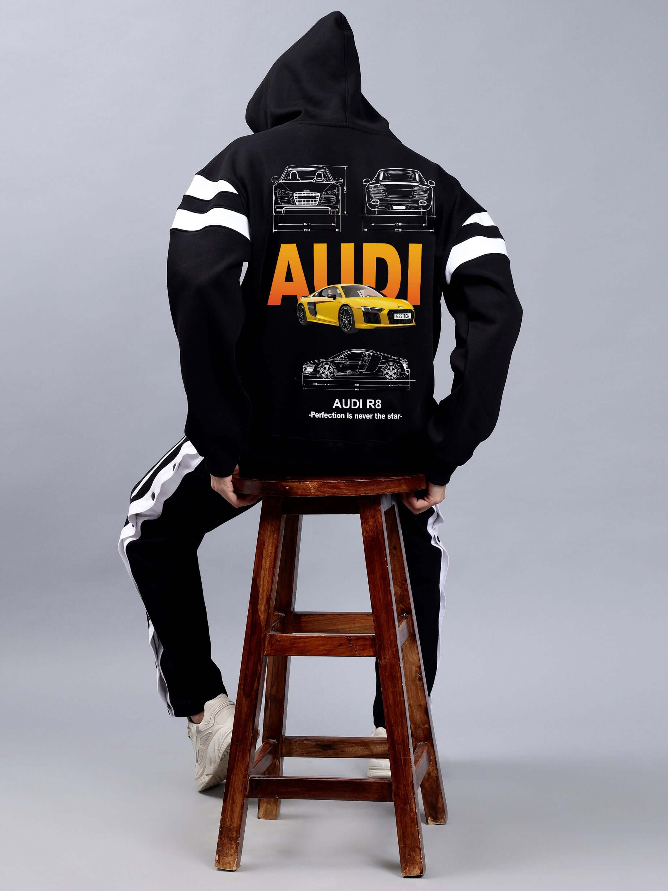 Back view of person wearing AUDI R8 FLEECE COLORBLOCK OVERSIZE HOODIE BLACK with car graphics, sitting on a stool against gray background.