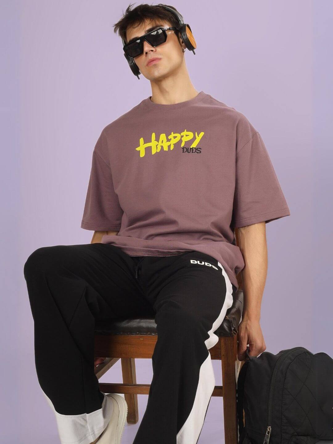Model wearing HAPPY DUDS OVER-SIZED T-SHIRT CHINESE VOILET with sunglasses and headphones, sitting on a chair.