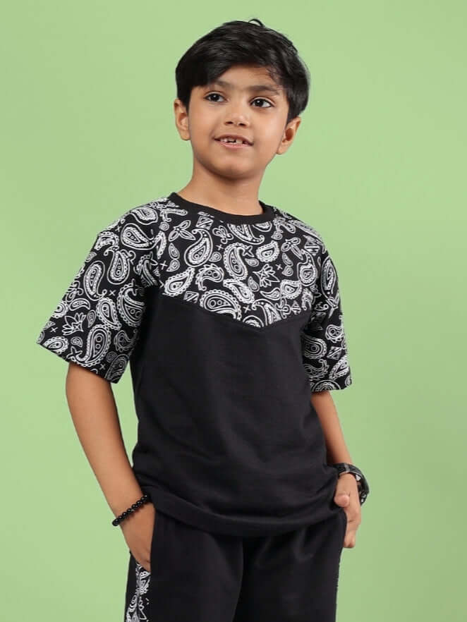 Young boy wearing a black Baladeer oversized T-shirt with paisley print for boys and girls, standing against a green background.