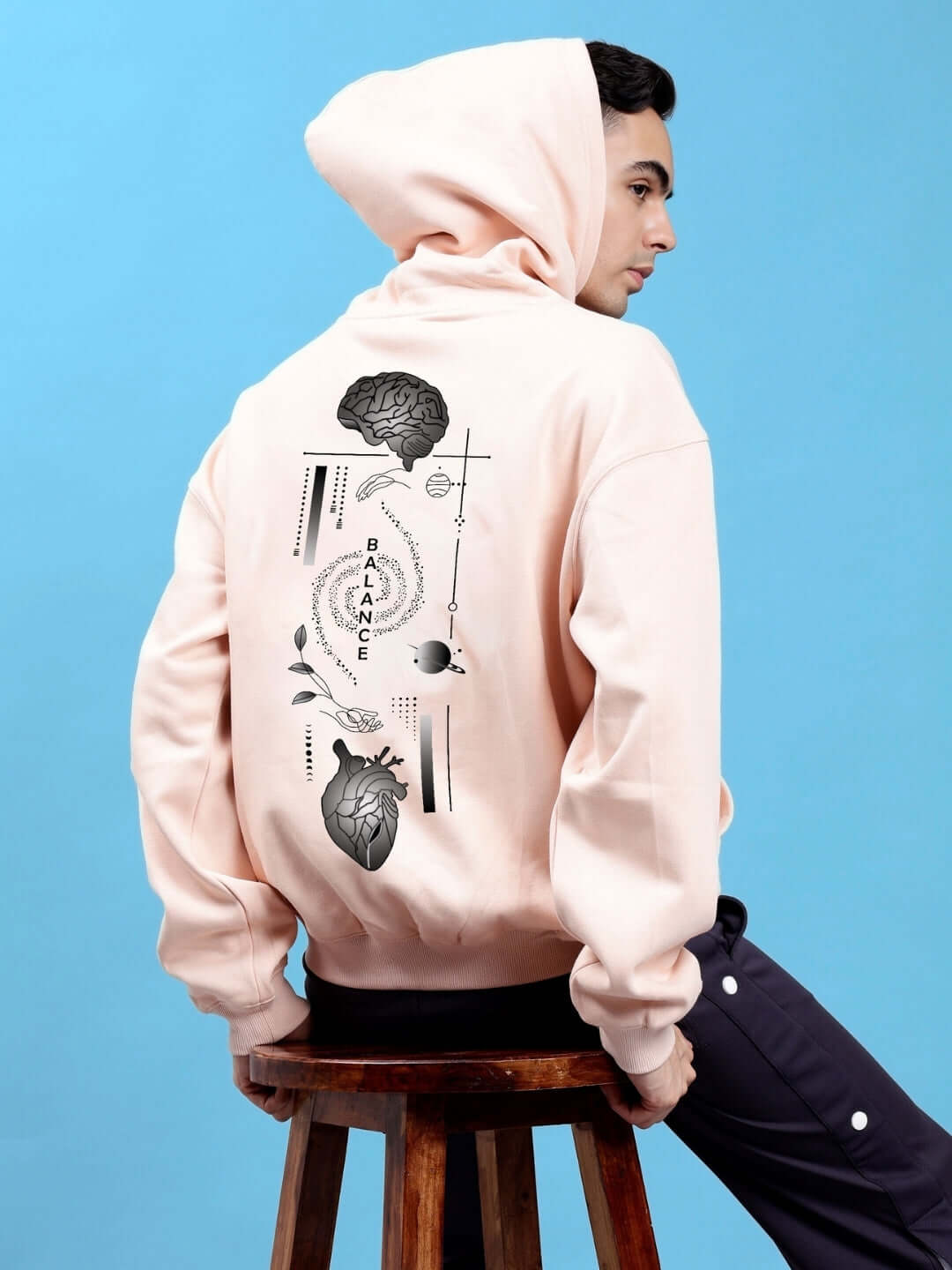 Man wearing BALANCE OVER-SIZED FLEECE HOODIES BEIGE with graphic back design, sitting on stool against blue background.