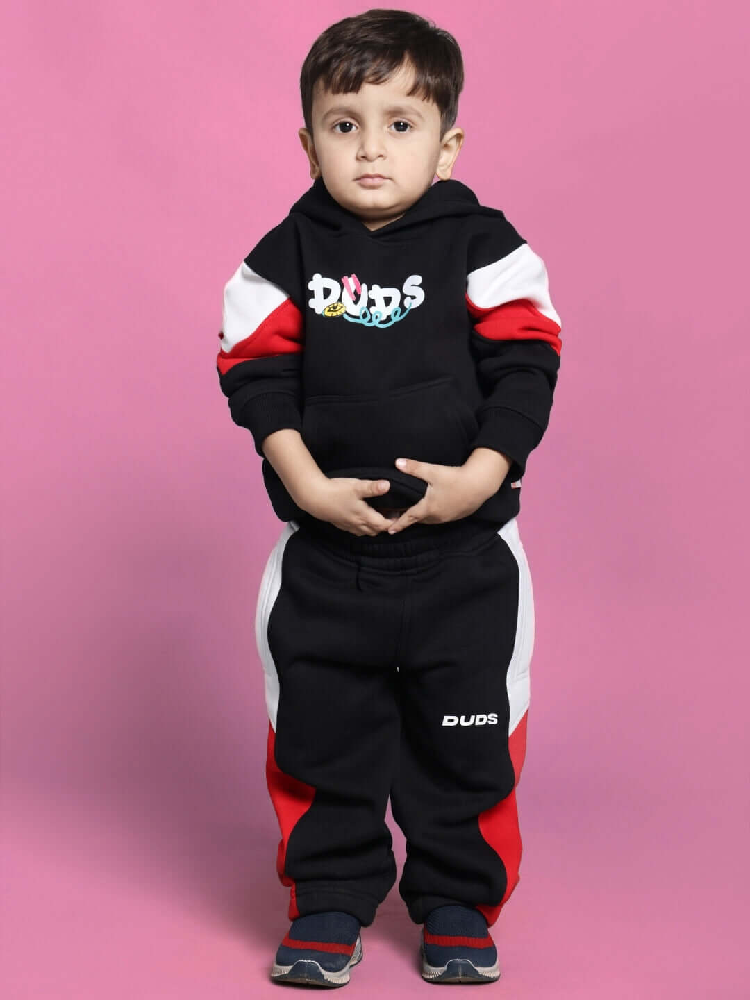 Child in black fleece beanie jogger suit with multicolor accents, standing against a pink background, showcasing casual kids fashion.