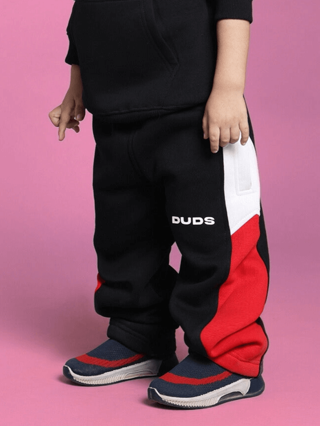 Child wearing black beanie fleece jogger pants with white and red accents and sneakers against a pink background.