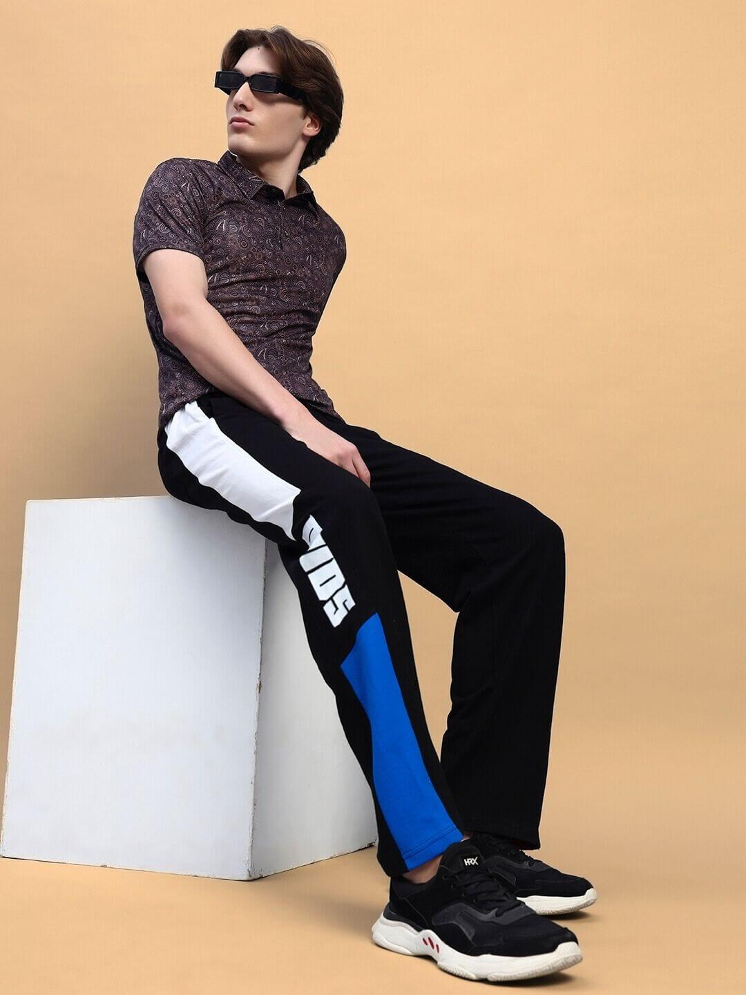 Model wearing BELGIANS RELAXED-FIT JOGGERS BLACK BLUE with black and blue design, seated stylishly against a beige backdrop.