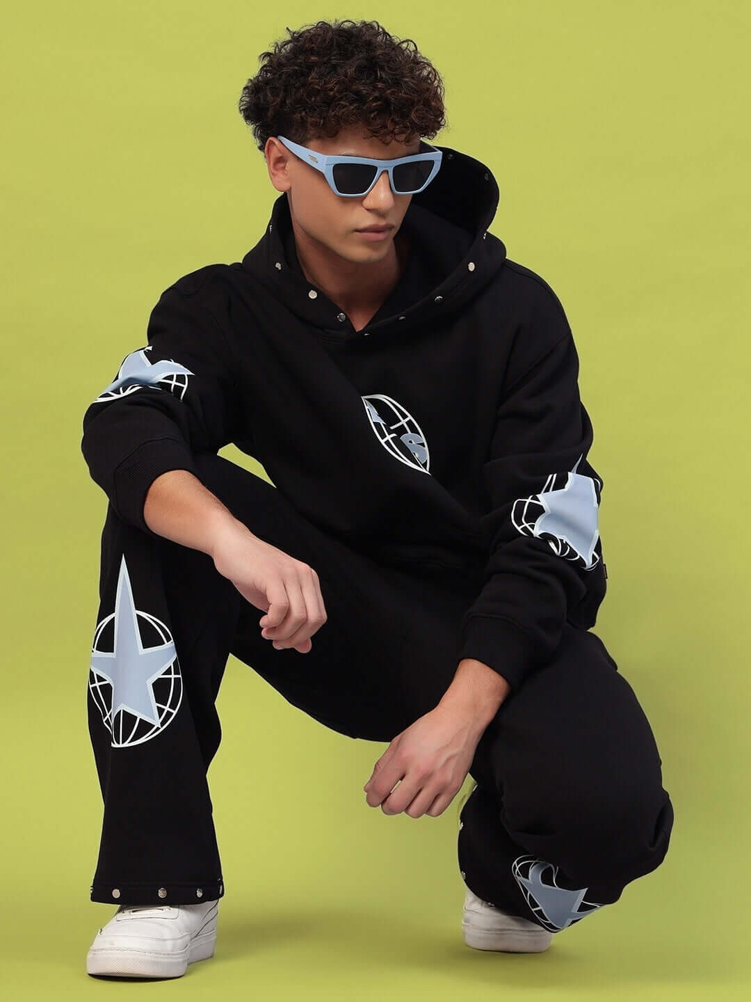 Model wearing Global Cotton Fleece Jogger in black with logo design, paired with sunglasses and sneakers against a green background.