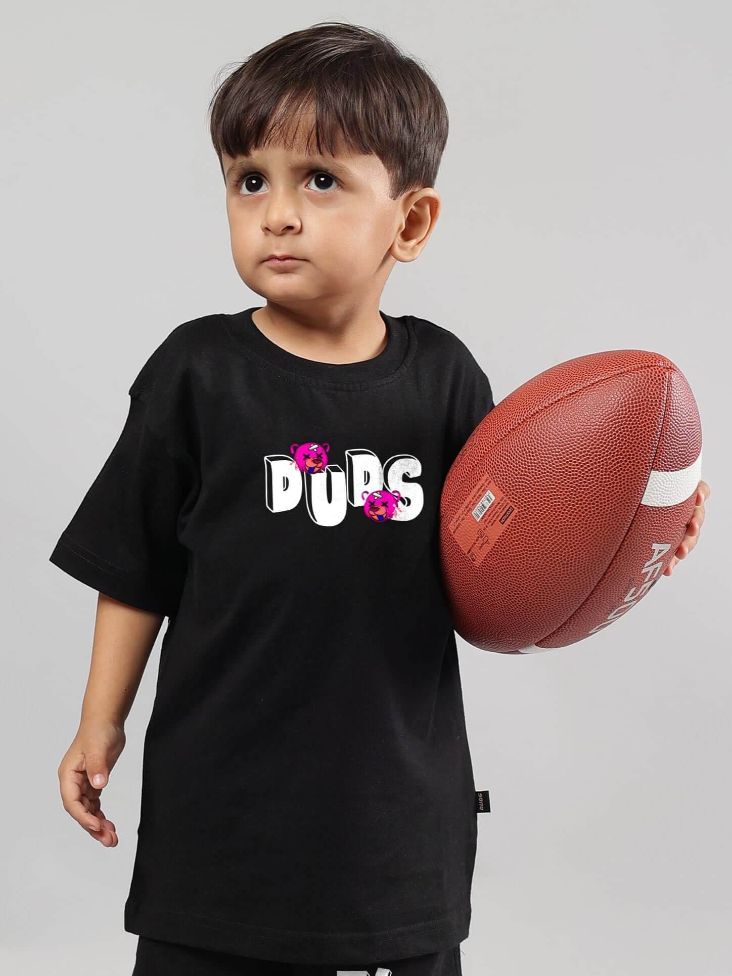 Young boy wearing a black I'M OK T-shirt holding a football, showcasing the stylish design by DUDS for kids. Perfect fit for boys and girls.