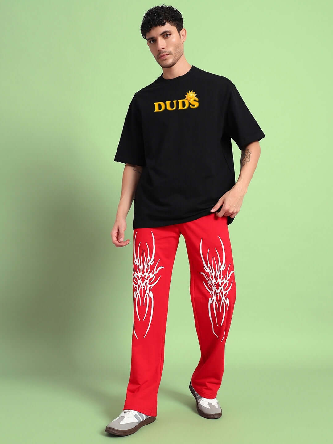 Man wearing a black t-shirt with 'DUDS' logo and red pants with white designs, standing in front of a green background.