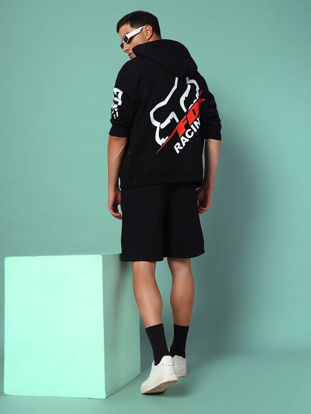 Model wearing RACING FOX FLEECE SHORTS BLACK with racing-inspired hoodie for a stylish streetwear look against a teal background.