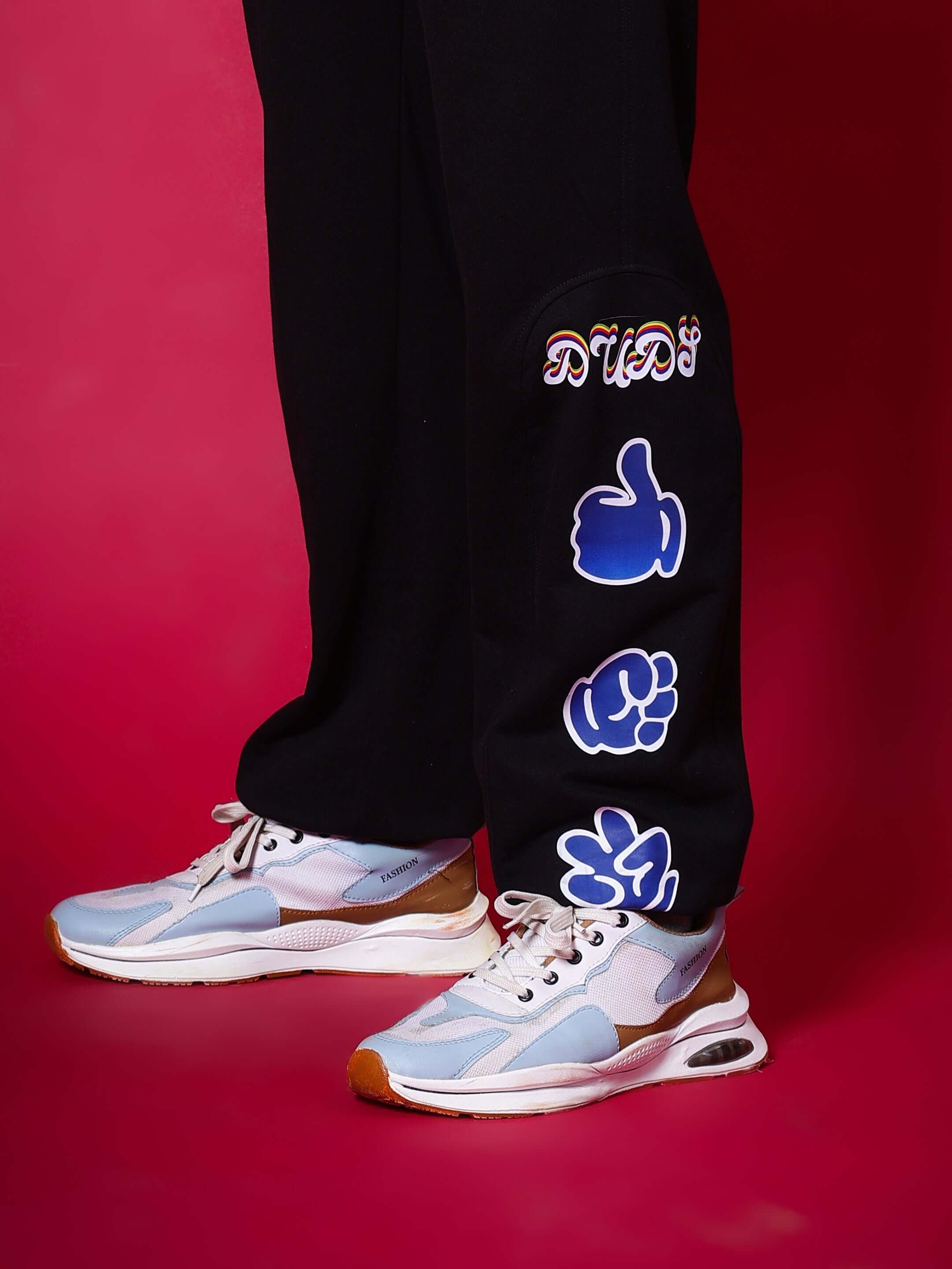 Black jogger pants featuring hand gesture designs by DUDS, worn with stylish sneakers, showcased on a vibrant red background.