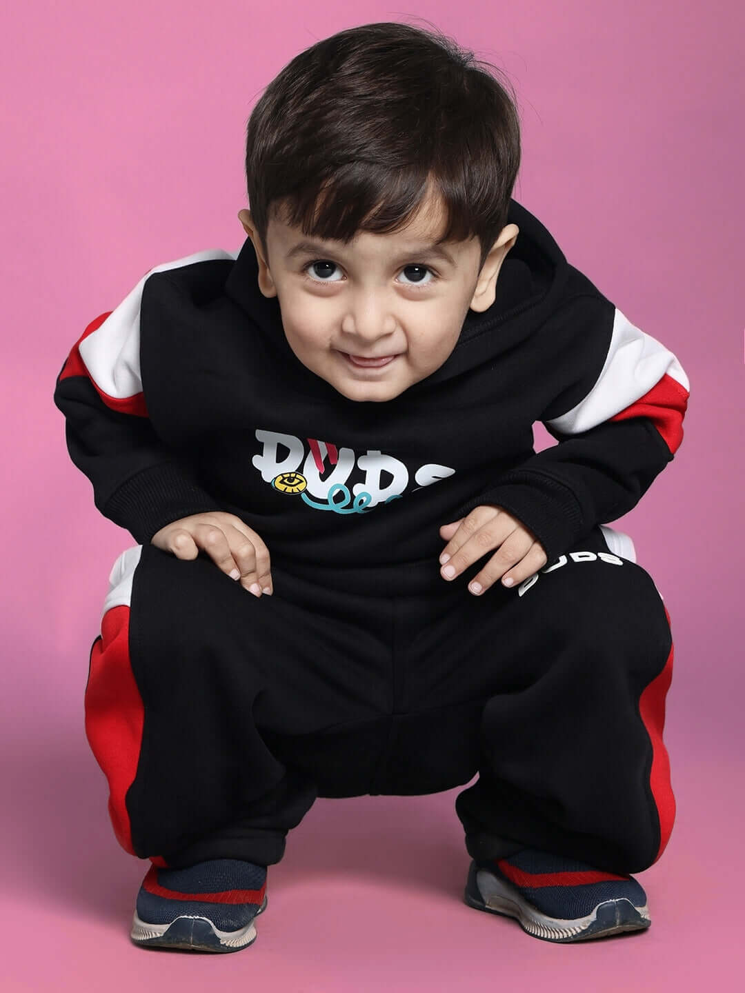 Child wearing black beanie hoodie by DUDS, with red and white accents, smiling against a pink background. Ideal for both boys and girls.