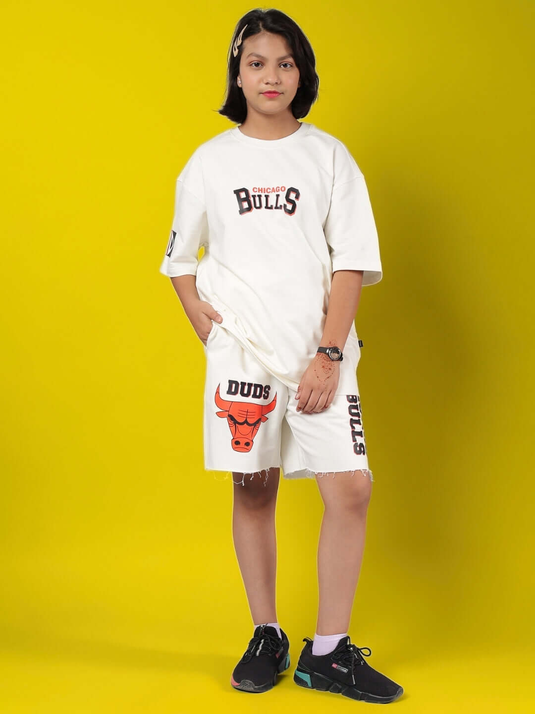 Child wearing BLAZE BULLS SHORTS FOR BOYS & GIRLS OFF-WHITE with Bulls branding, standing against a yellow background.