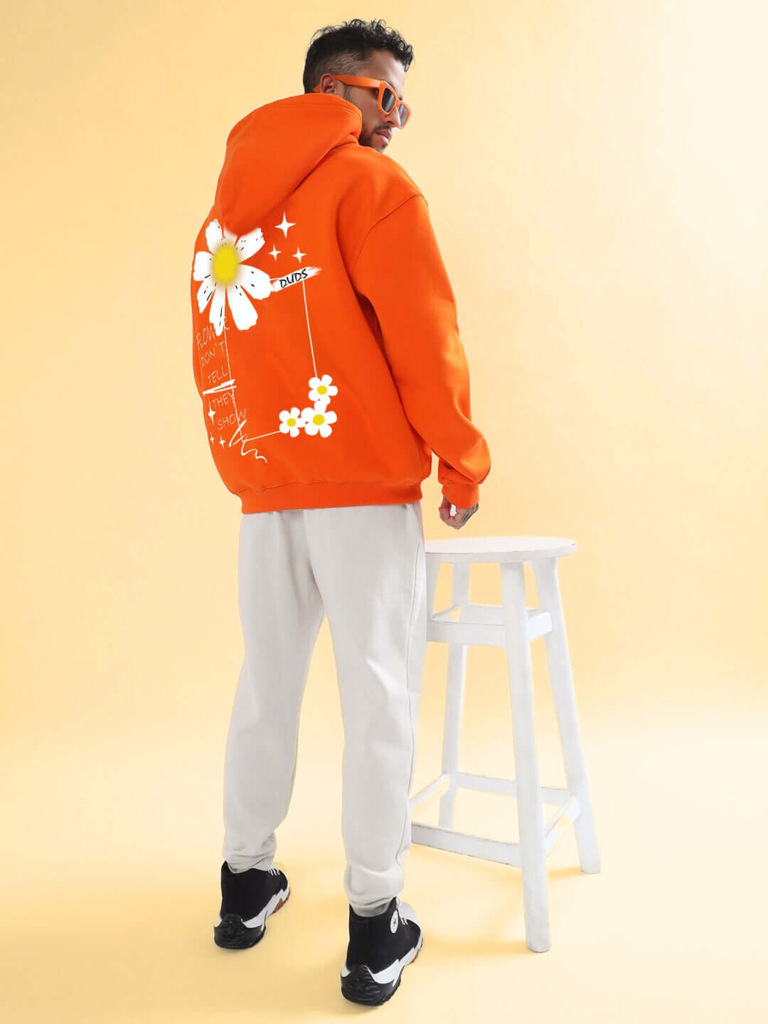 Man wearing BLOOM QUOTE FLEECE OVERSIZED HOODIE ORANGE with floral back design, styled with light pants and sneakers.