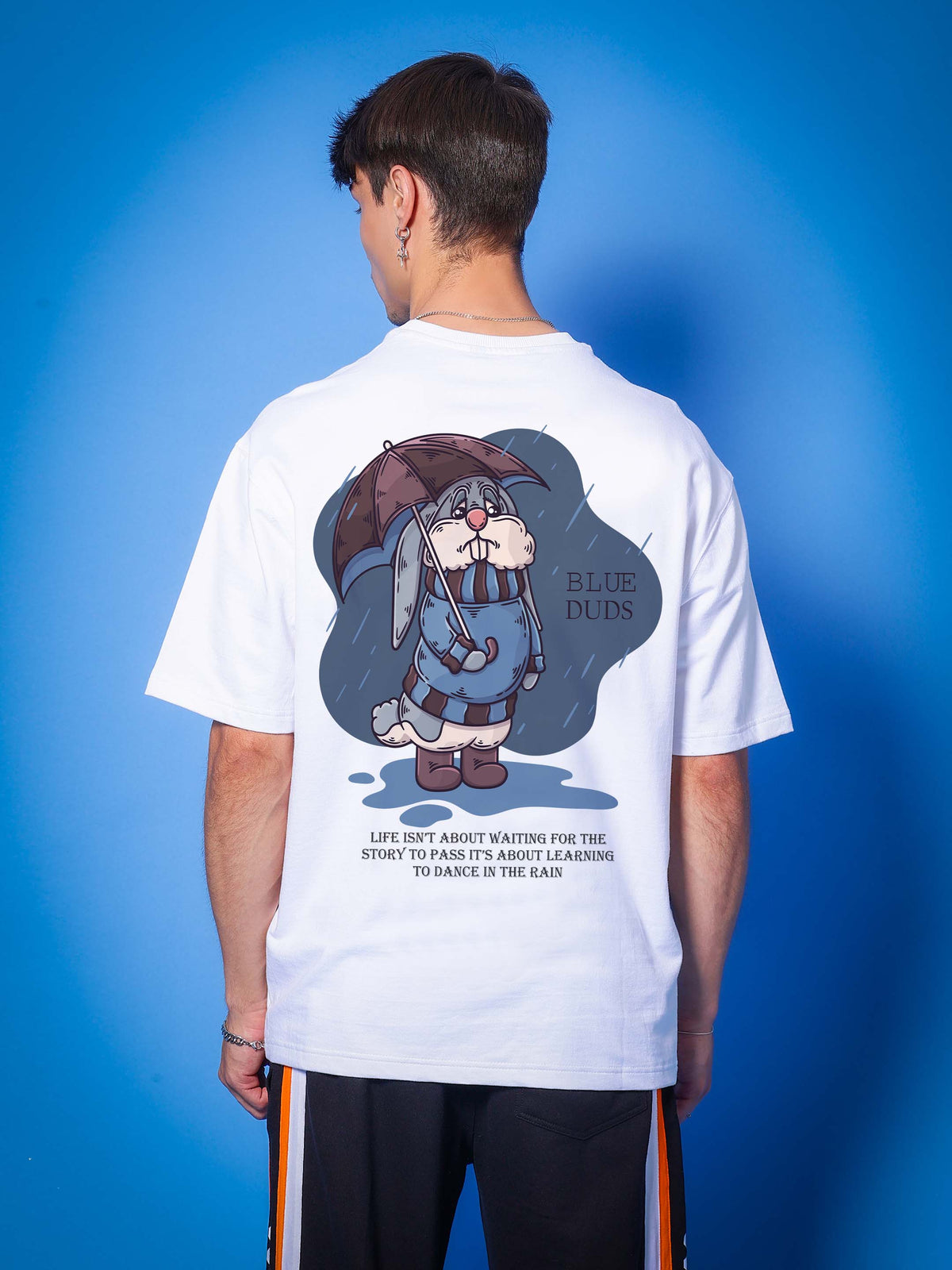 Man wearing BLUE LAPIN OVER-SIZED T-SHIRT WHITE with cartoon graphic and inspirational quote on the back, blue background.