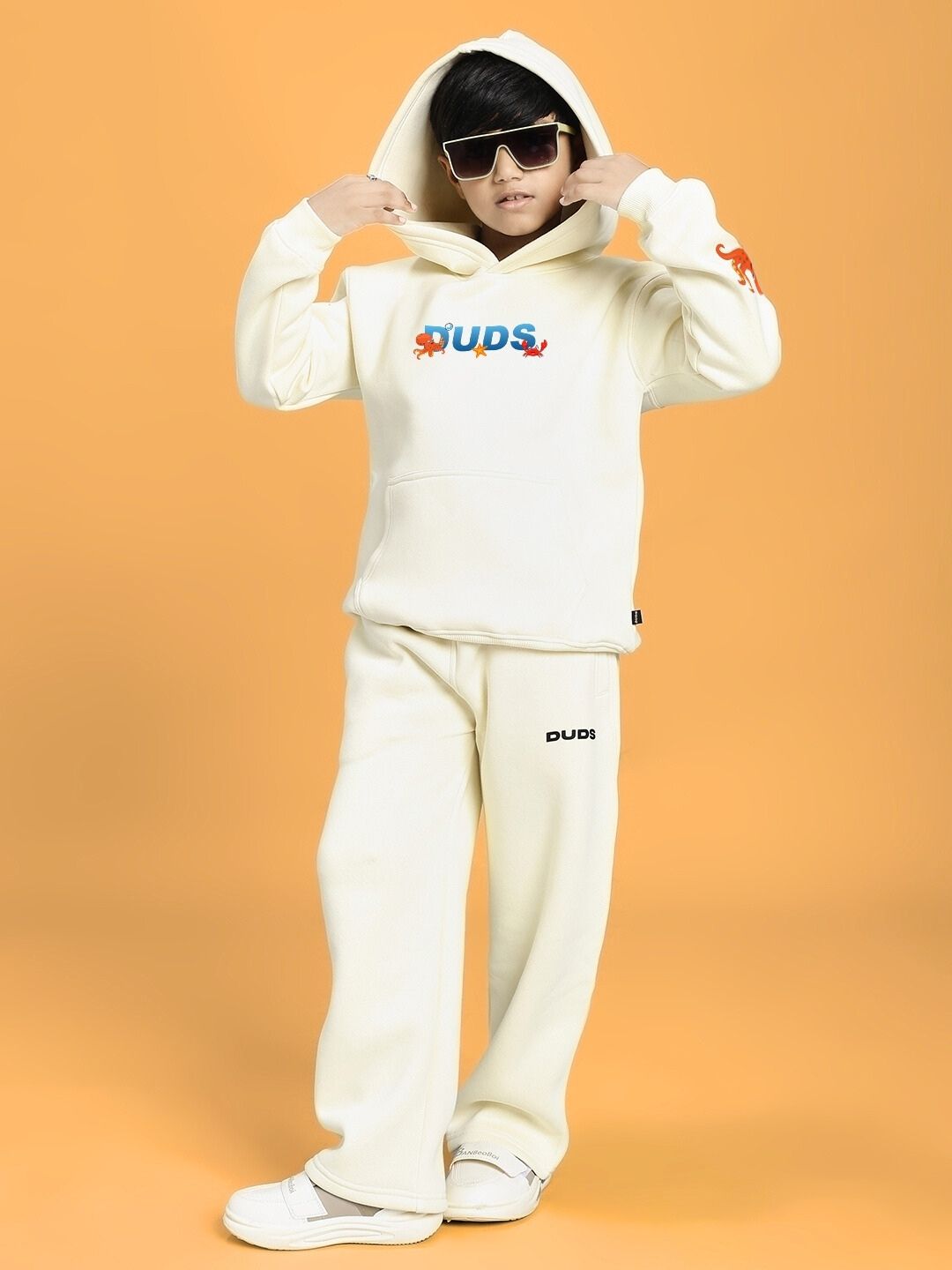 Child wearing BLUE OCEAN WORLD FLEECE JOGGER FOR BOYS AND GIRLS OFF-WHITE with a hood up, against an orange background.