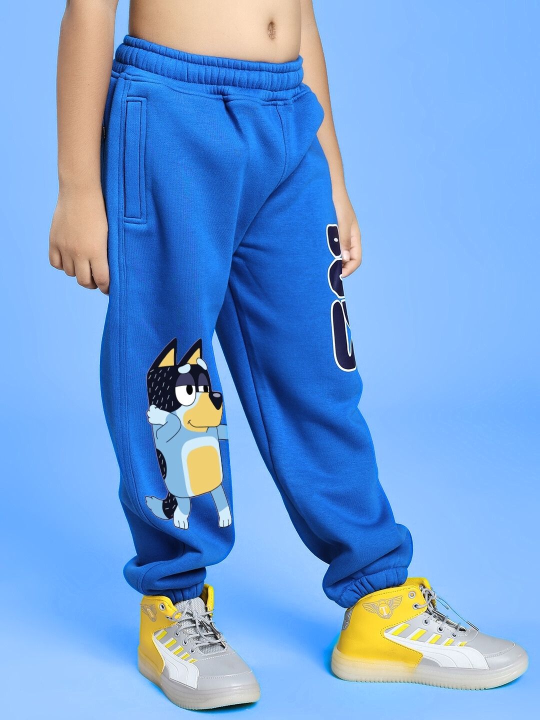 Child wearing BLUEY BINGO FLEECE JOGGER FOR BOYS AND GIRLS BLUE with cartoon character print on blue pants, standing on blue background.