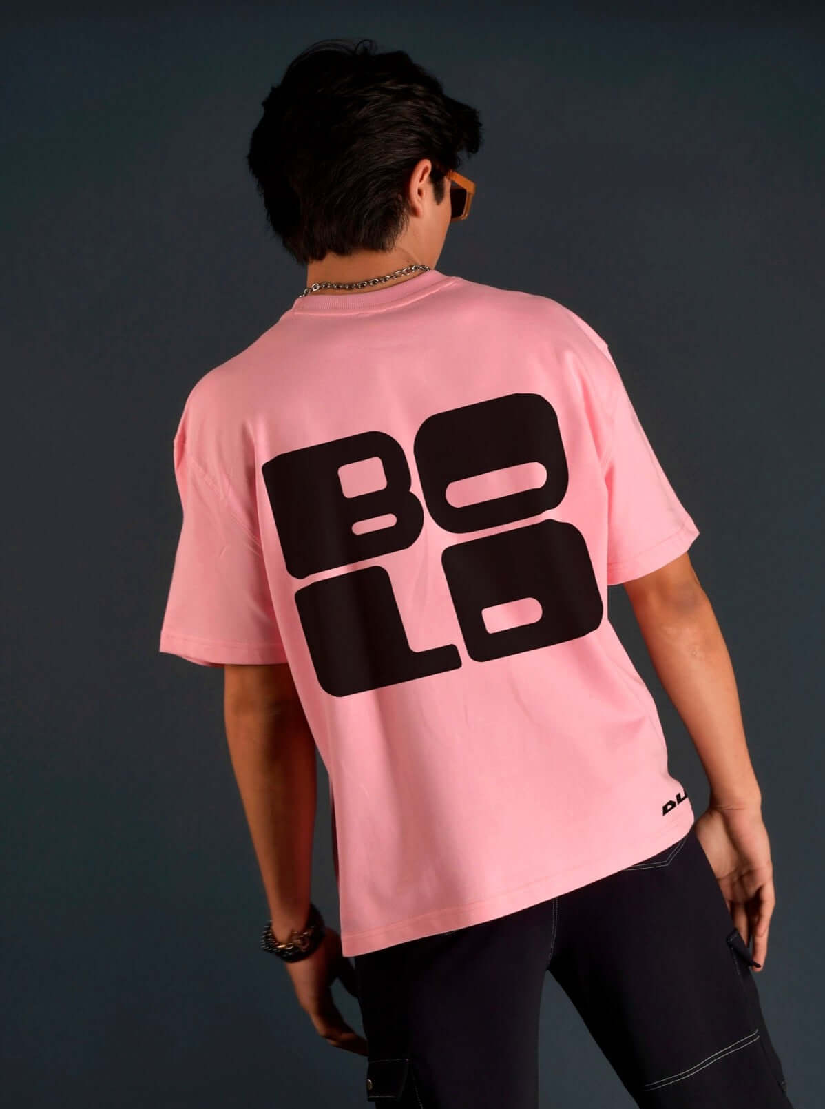 Man wearing a baby pink over-sized t-shirt with bold black 'BOLO' design on the back, standing against a dark background.
