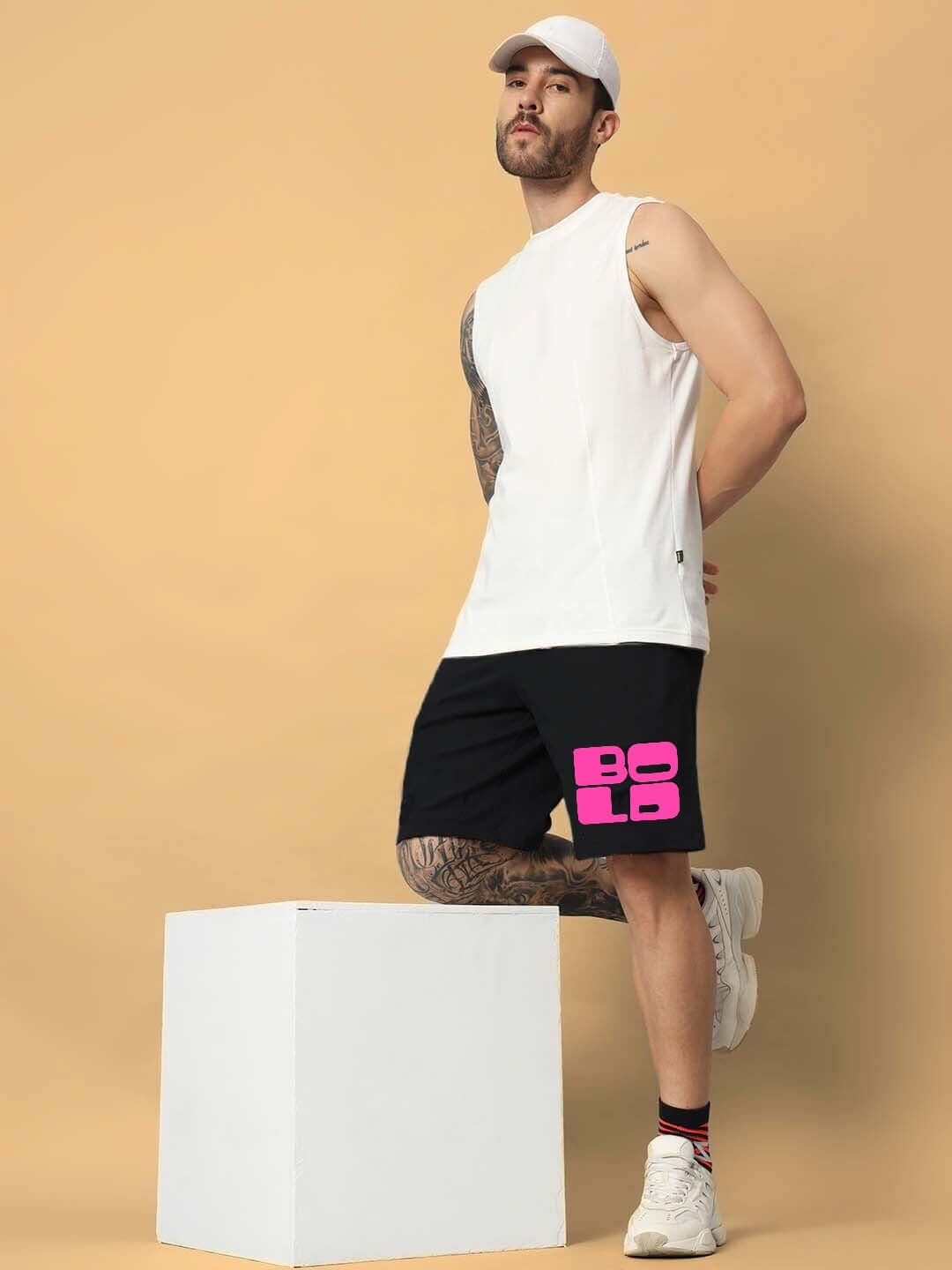 Man wearing BOLD Regular Fit Black Shorts and white tank top, leaning on a white box, showcasing casual fashion ensemble.