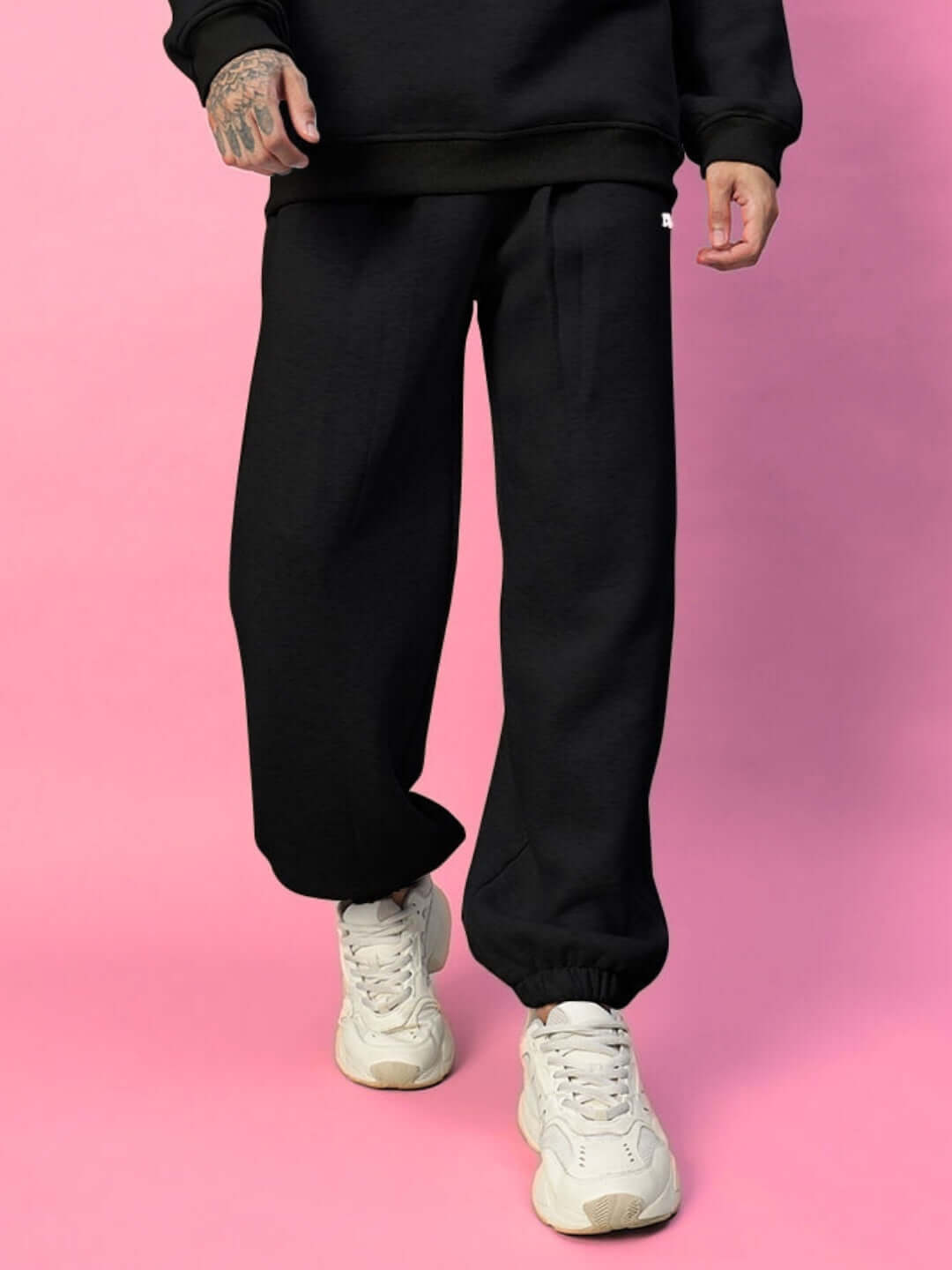 Model wearing BONA-FIDE Cotton Fleece Pant in Black, showcasing relaxed fit and elastic cuffs, paired with white sneakers against a pink background.