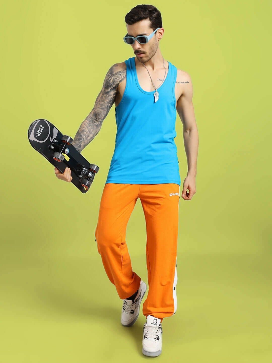 Man in bright blue gym t-shirt, holding a skateboard, wearing orange pants and sunglasses, stands against a green background.