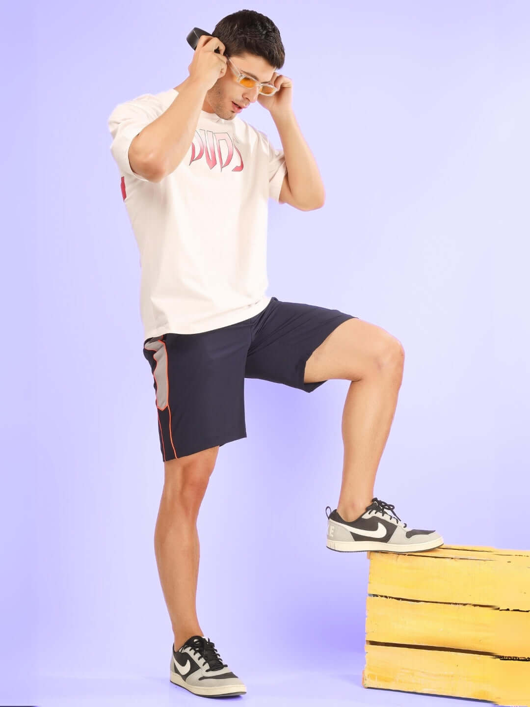Man wearing BOOMER SHORTS NAVY WITH ORANGE PIPING and sneakers, posing on a box in a stylish streetwear outfit.