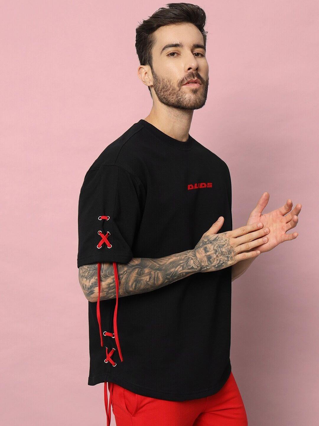 Man wearing BOWLINE OVER-SIZED T-SHIRT BLACK with red accents, standing against a pink background.