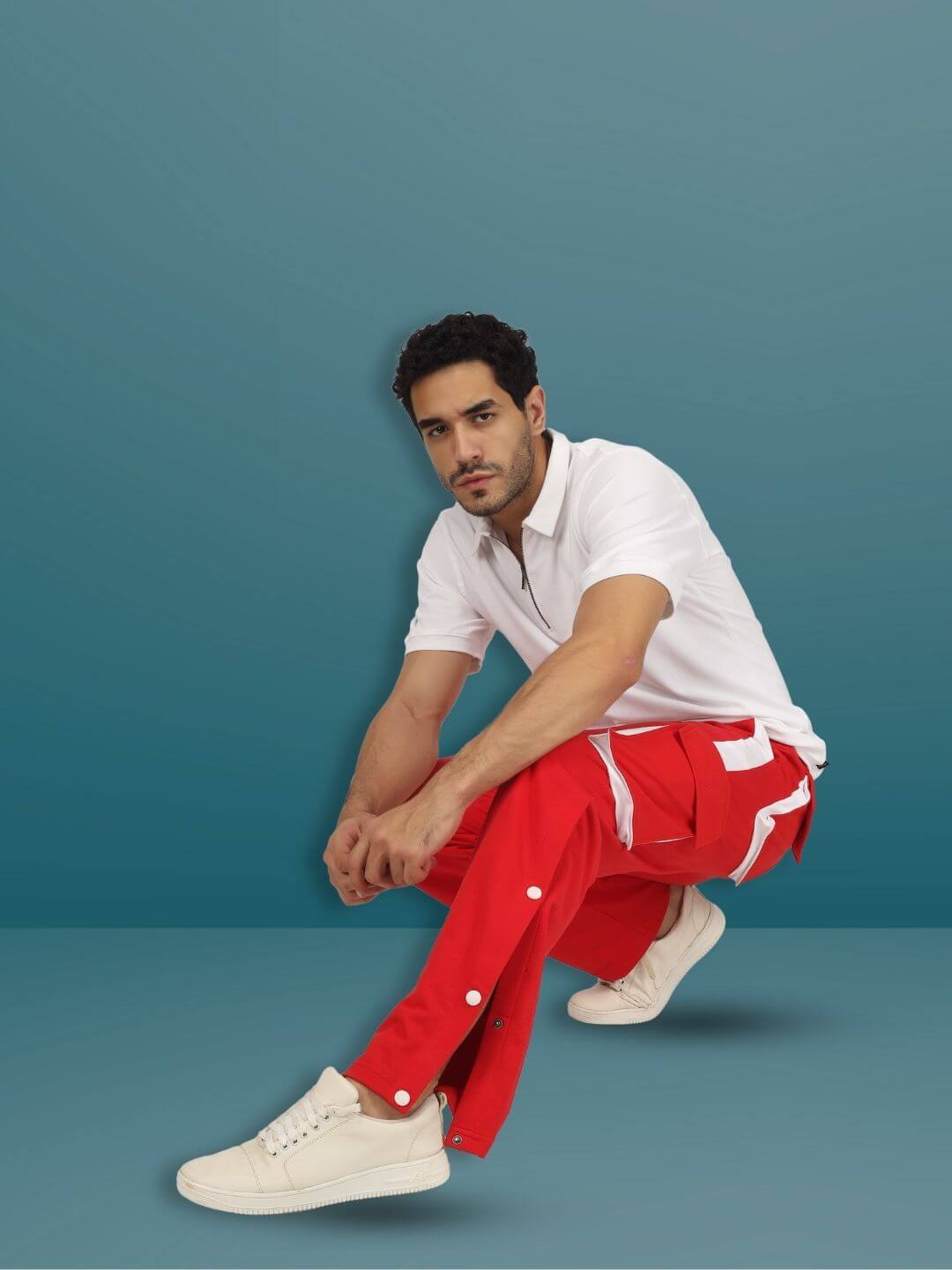 Man wearing BRACKET CARGO PANT in red with white highlights, crouching on a blue background, showcasing casual style.