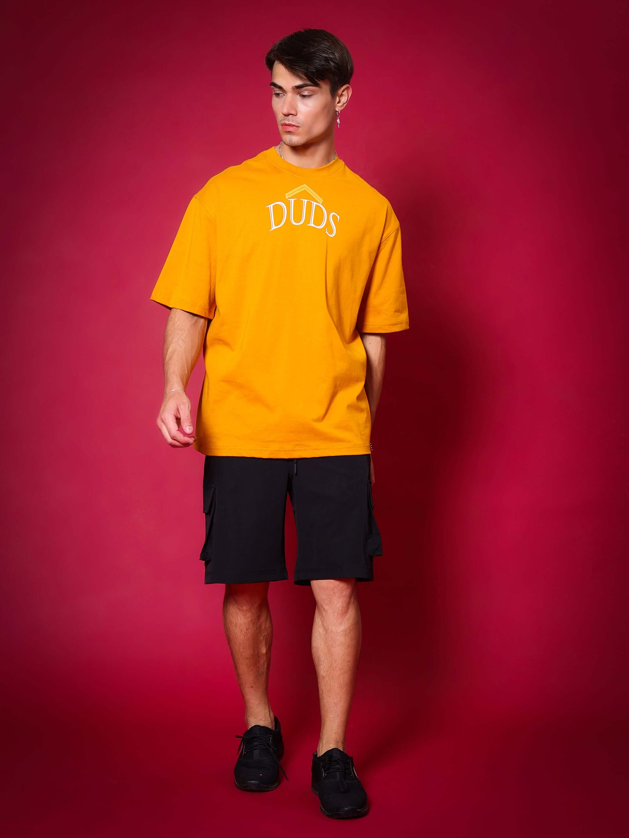 Model wearing black Brawny Cargo Shorts with yellow DUDS T-shirt, showcasing custom fit option against a red background.