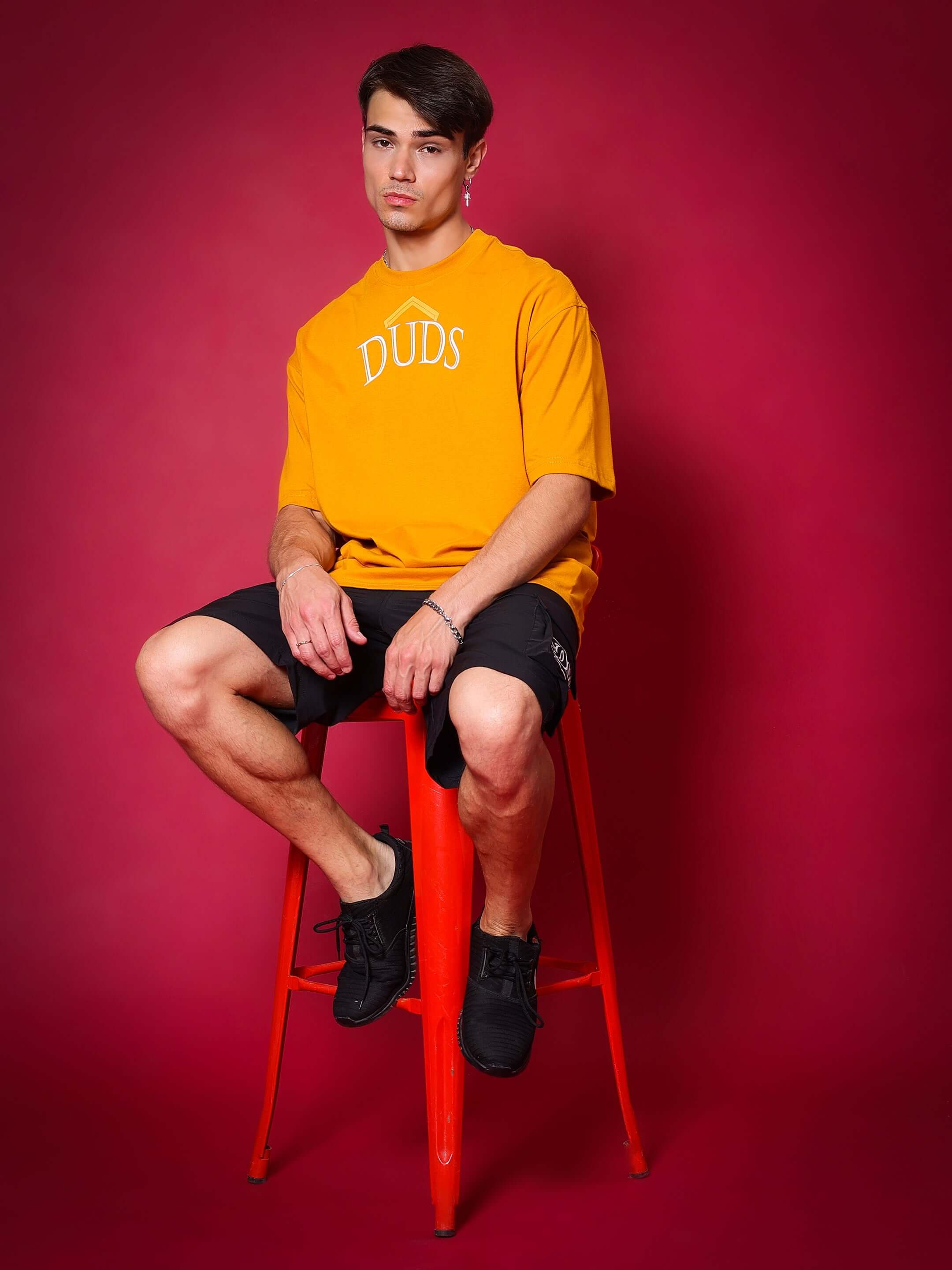 Man wearing orange DUDS shirt and black BRAWNY cargo shorts sitting on red stool against a red background.