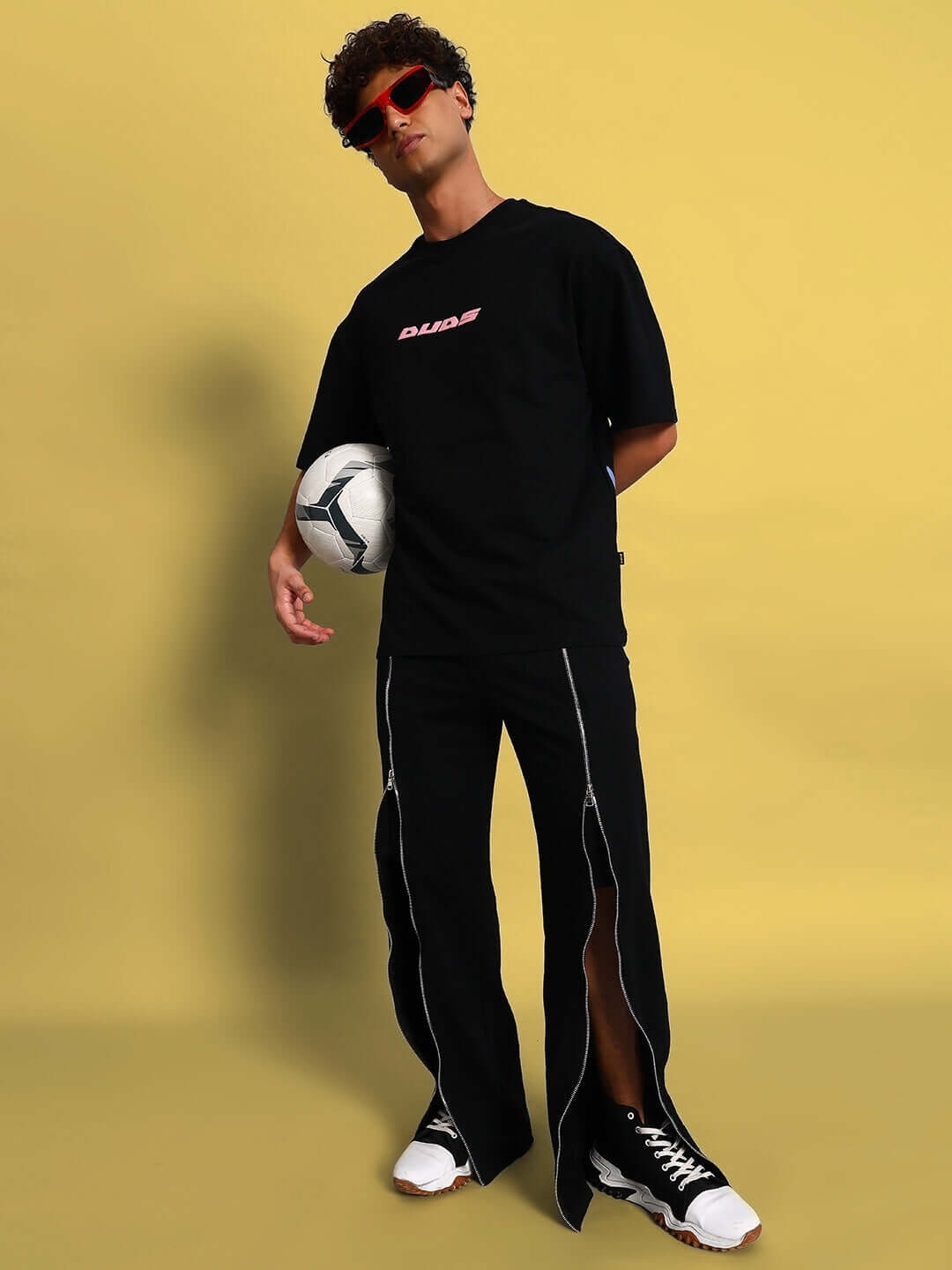 Model wearing black Brazo front open zipper joggers, holding a soccer ball, against a yellow background.