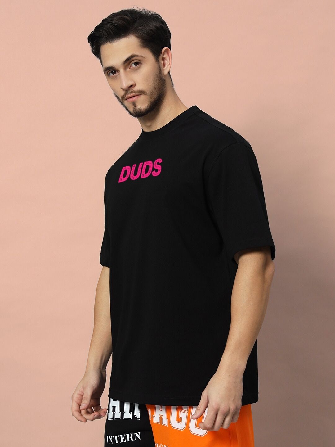 Man wearing BREAKING OVER-SIZED T-SHIRT BLACK with pink lettering against a beige background.