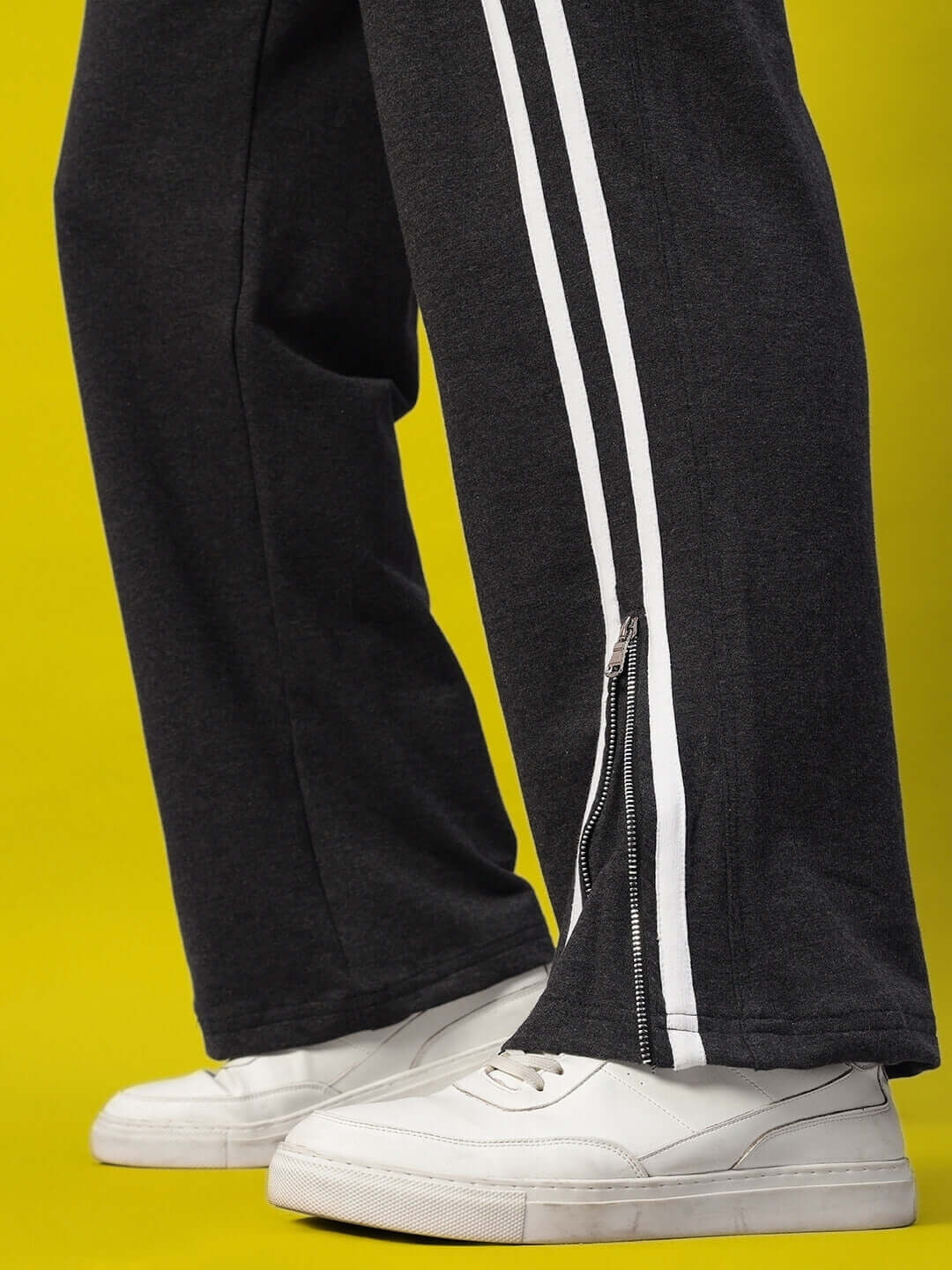 Dark grey BRONCO slit zipper joggers with white stripes, paired with white sneakers, against a yellow background.