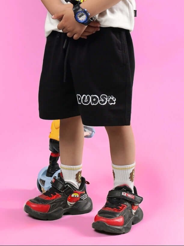 Child wearing BRUNO SHORTS FOR BOYS & GIRLS BLACK by DUDS with colorful sneakers on a pink background