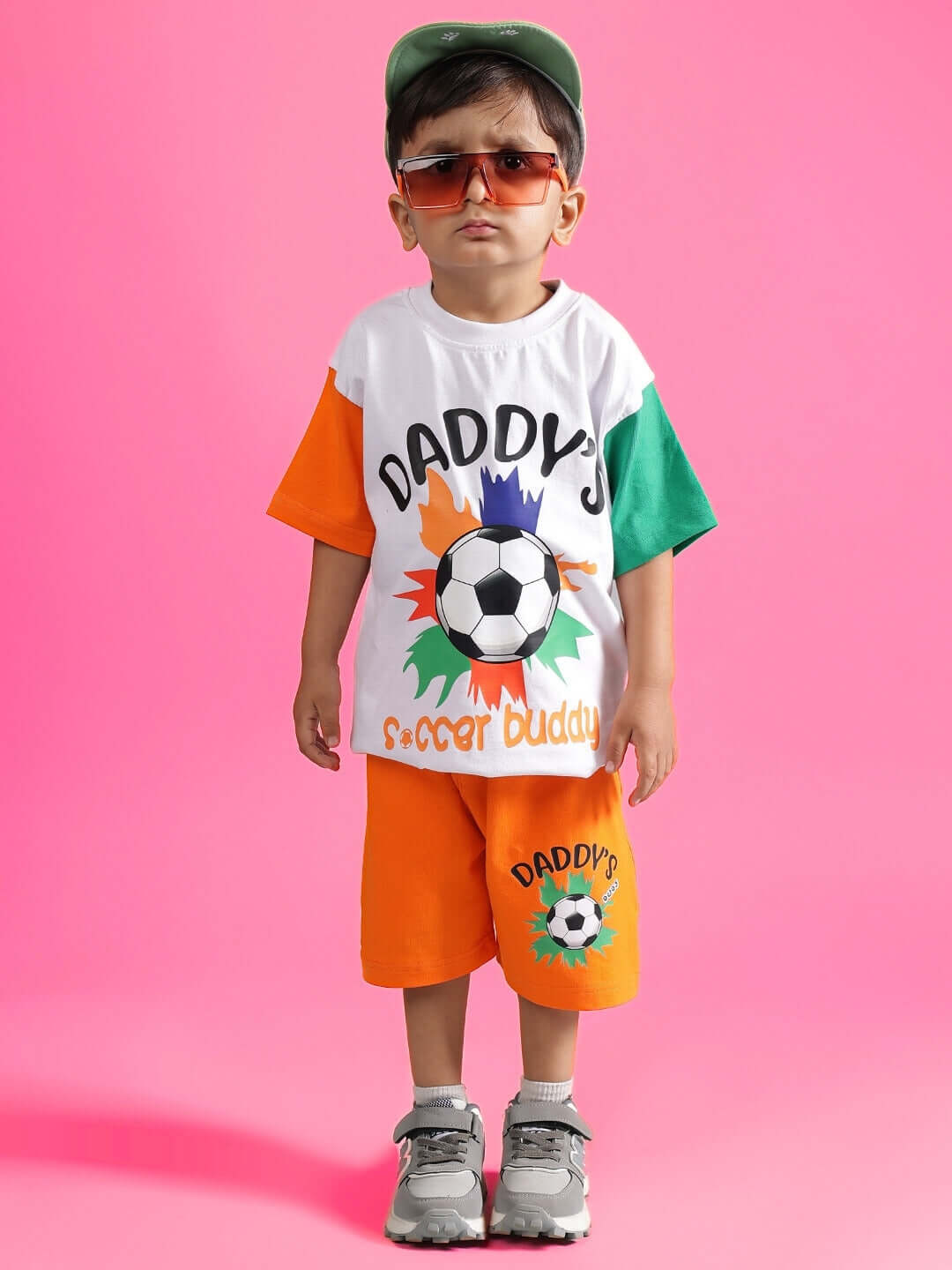 Child wearing Buddy Co-ord set in white and orange with soccer design, standing against a pink background. Ideal for boys and girls.