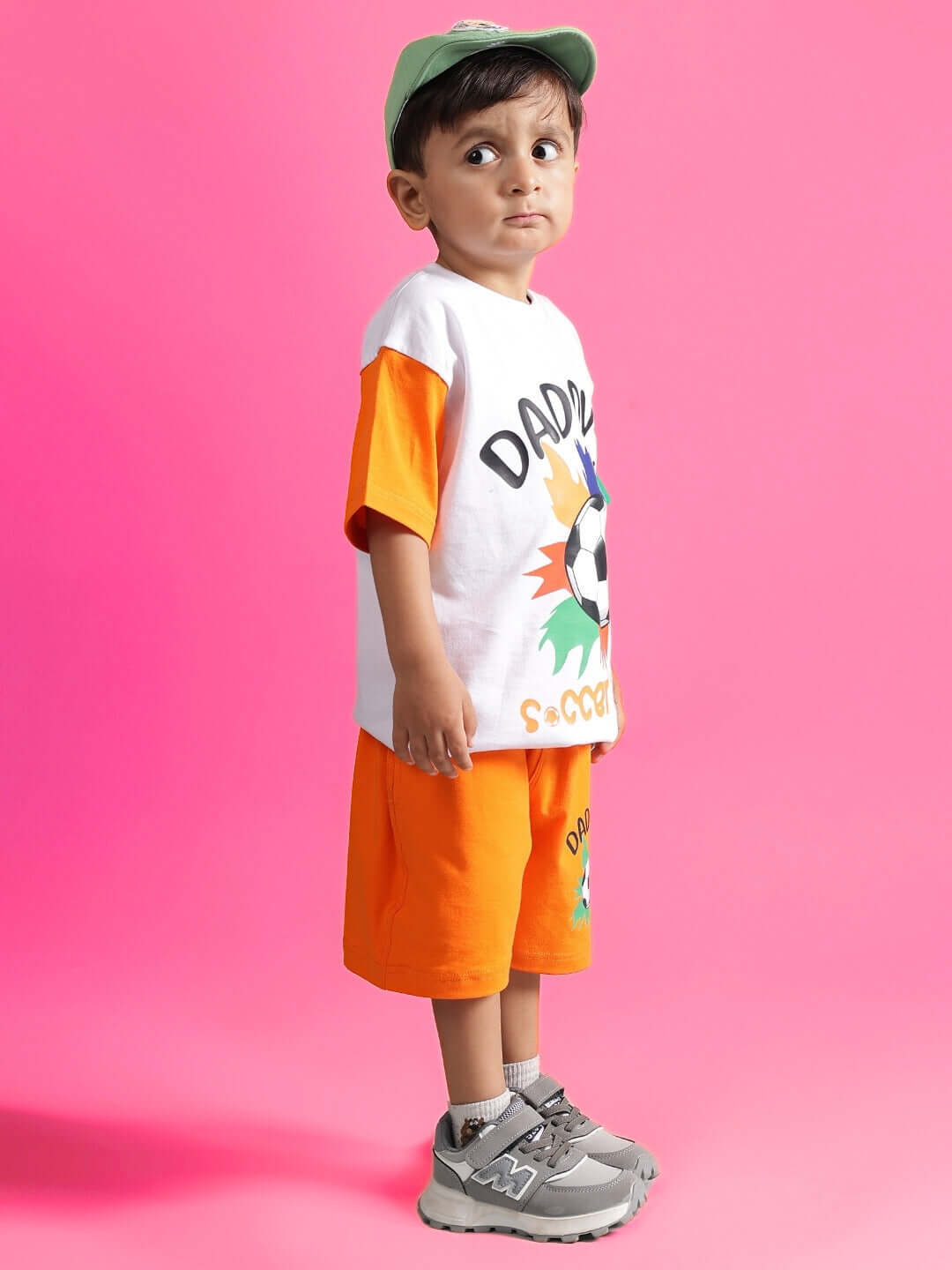Child wearing BUDDY CO-ORD FOR BOYS & GIRLS WHITE-ORANGE outfit with cap and sneakers against a pink background