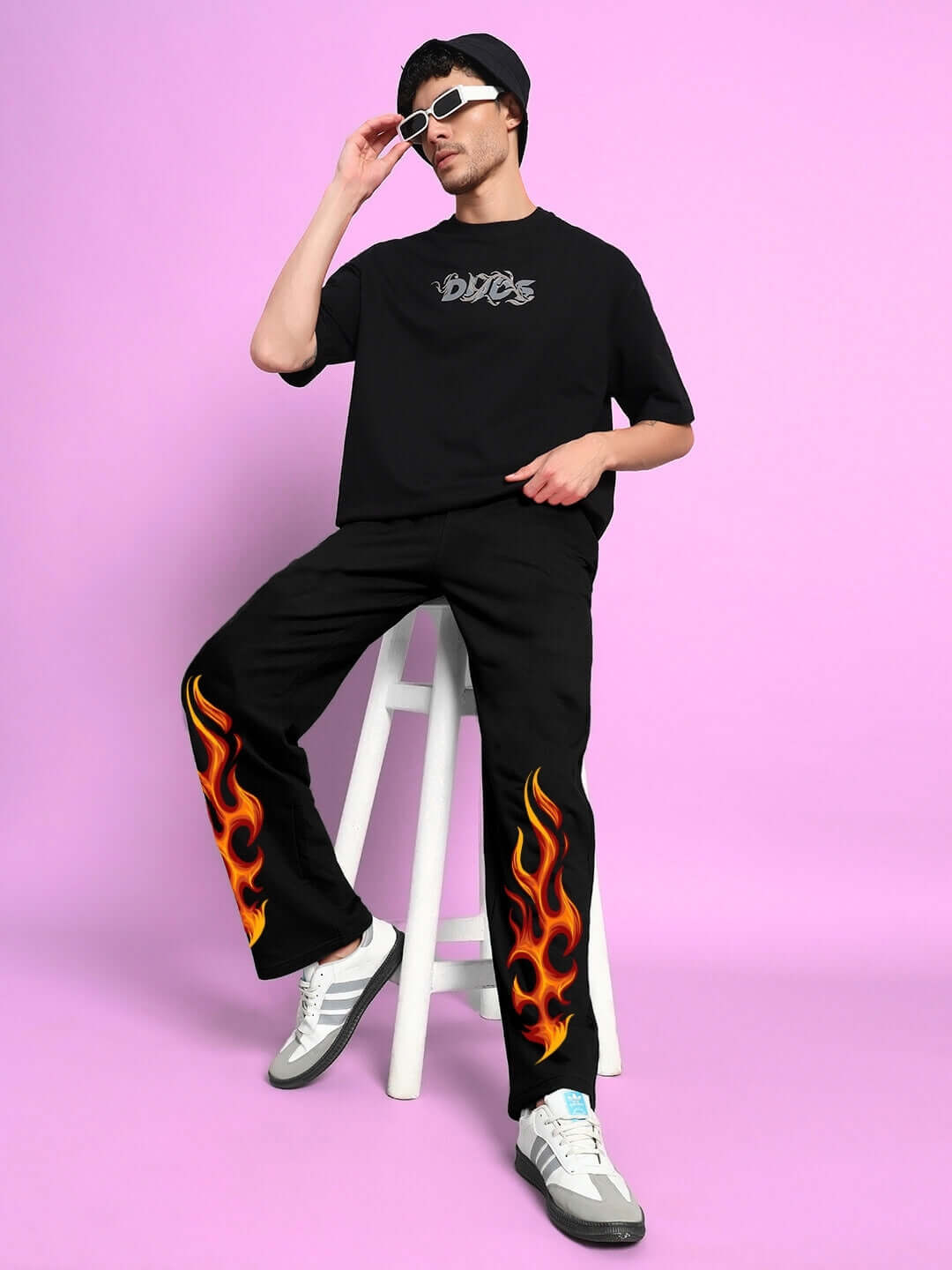 Model wearing black Burning Fire Jogger with flame design, sitting on a stool against a pink background, showcasing trendy DUDS apparel.