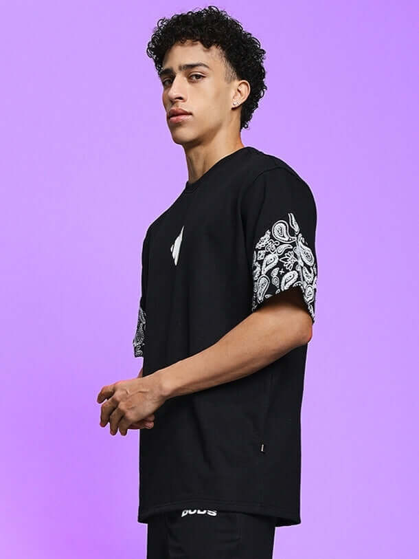 Model wearing CALVIN black oversized t-shirt with paisley sleeves, premium cotton blend, ideal for casual wear, against purple background.