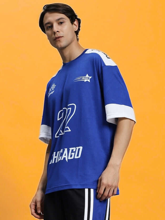 Model wearing CHALLENGE OVER-SIZED T-SHIRT ROYAL BLUE with graphics, showcasing casual style against orange background.