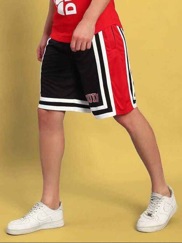 Model wearing red and black Challenge Regular Fit Shorts with white sneakers on a yellow background.