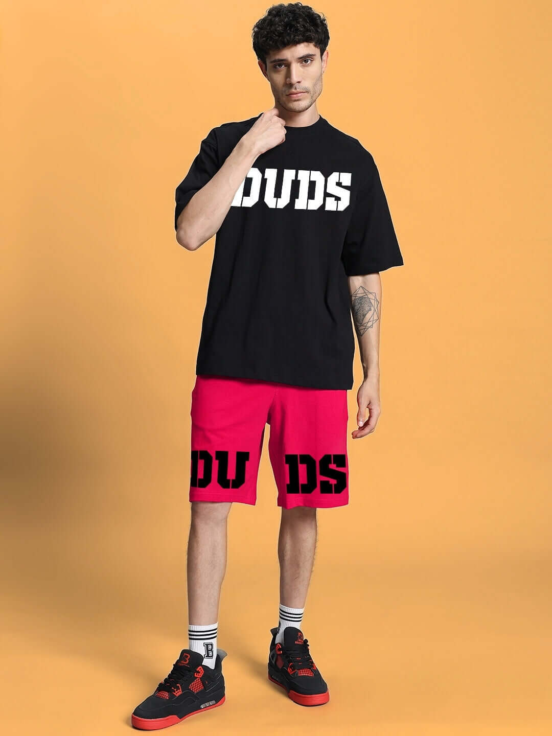 Model wearing black DUDS T-shirt and pink shorts with bold black logo, stylish casual outfit, orange background for contrast.