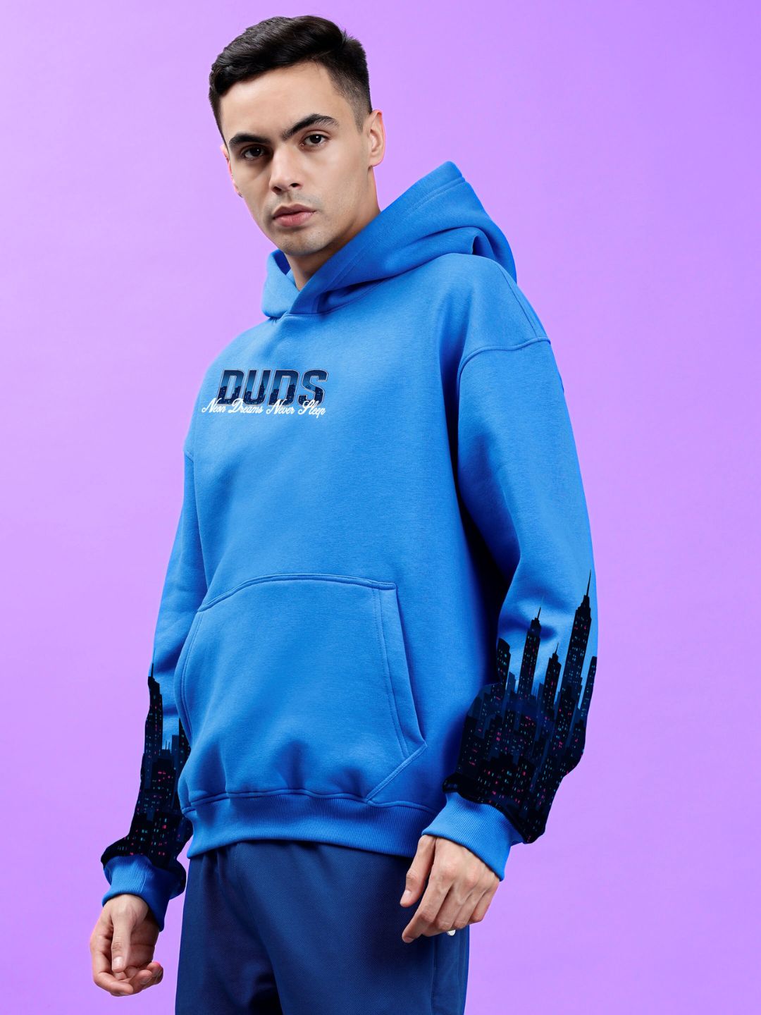 Model wearing CITY OF DREAMS PRINTED HOODIE ROYAL BLUE, showcasing bold color and street-style design on a purple backdrop.