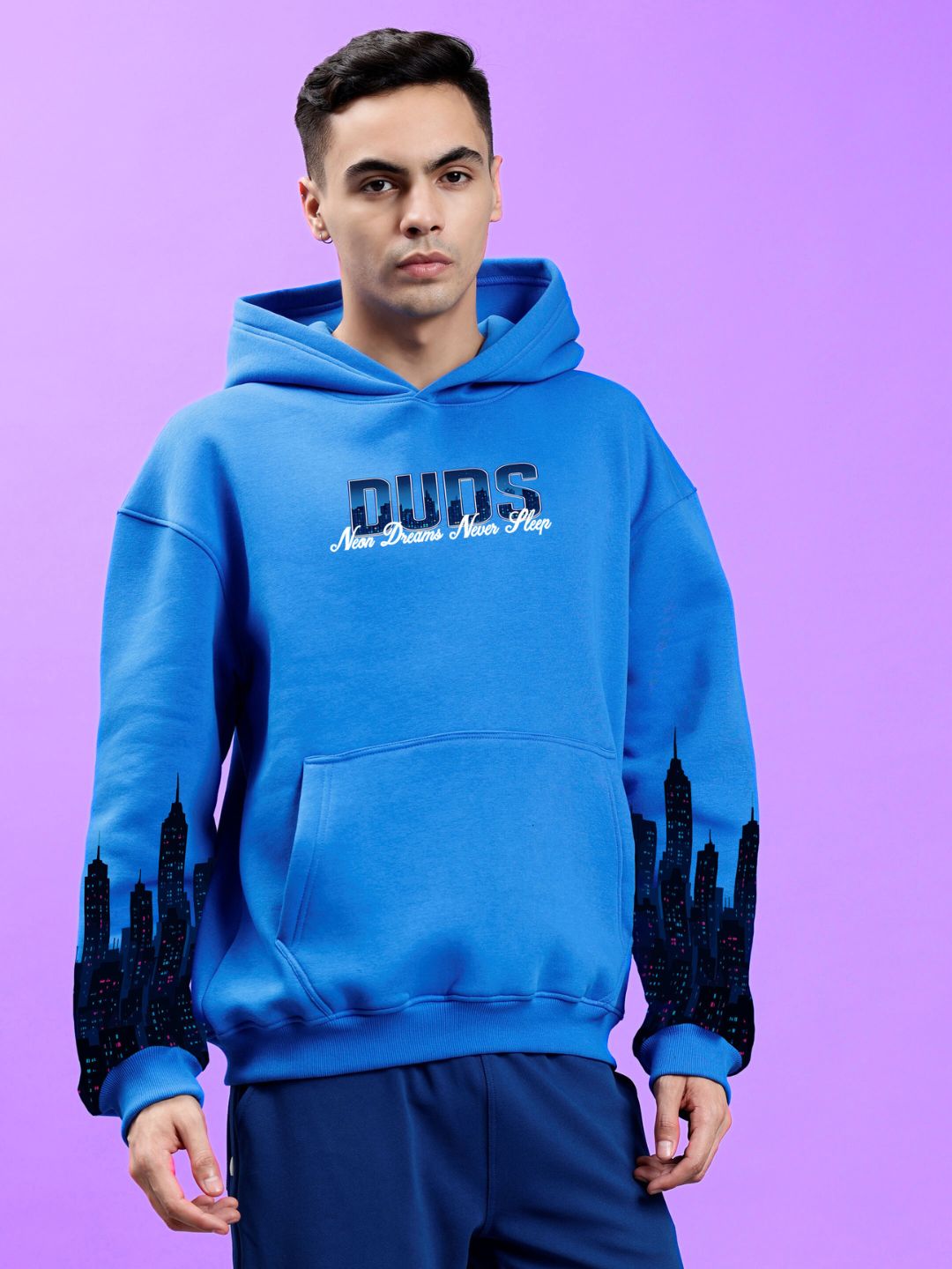 Model wearing CITY OF DREAMS PRINTED HOODIE ROYAL BLUE with city skyline print against a purple background
