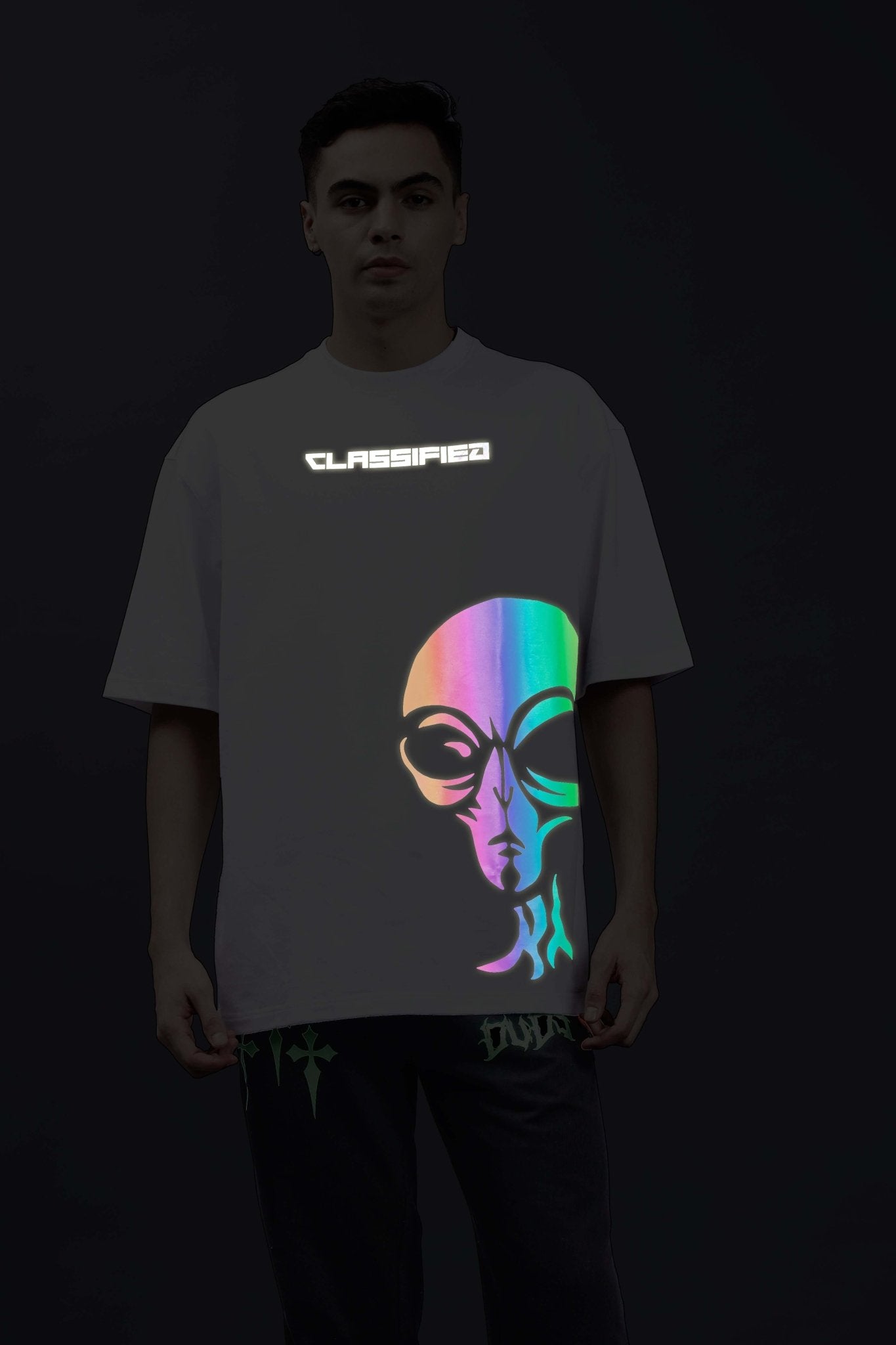 Model wearing CLASSIFIED REFLECTOR OVER-SIZED T-SHIRT WHITE with reflective alien graphic and logo in a dark setting.