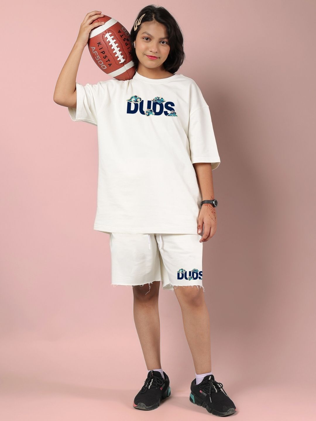 Child wearing CLOUD PRINTED SHORTS FOR BOYS AND GIRLS OFF WHITE, holding football, standing against pink background.