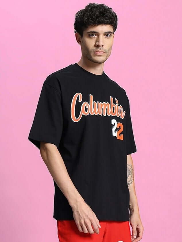 Person wearing COLUMBIA 22 PRINT OVER-SIZED T-SHIRT BLACK against pink background, showcasing bold graphic design and casual style.