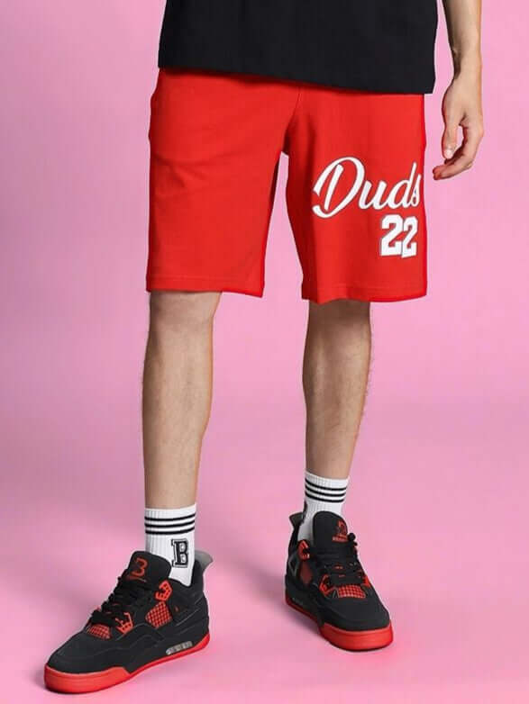 COLUMBIA 22 REGULAR FIT SHORTS RED modeled with sneakers and socks on a pink background.