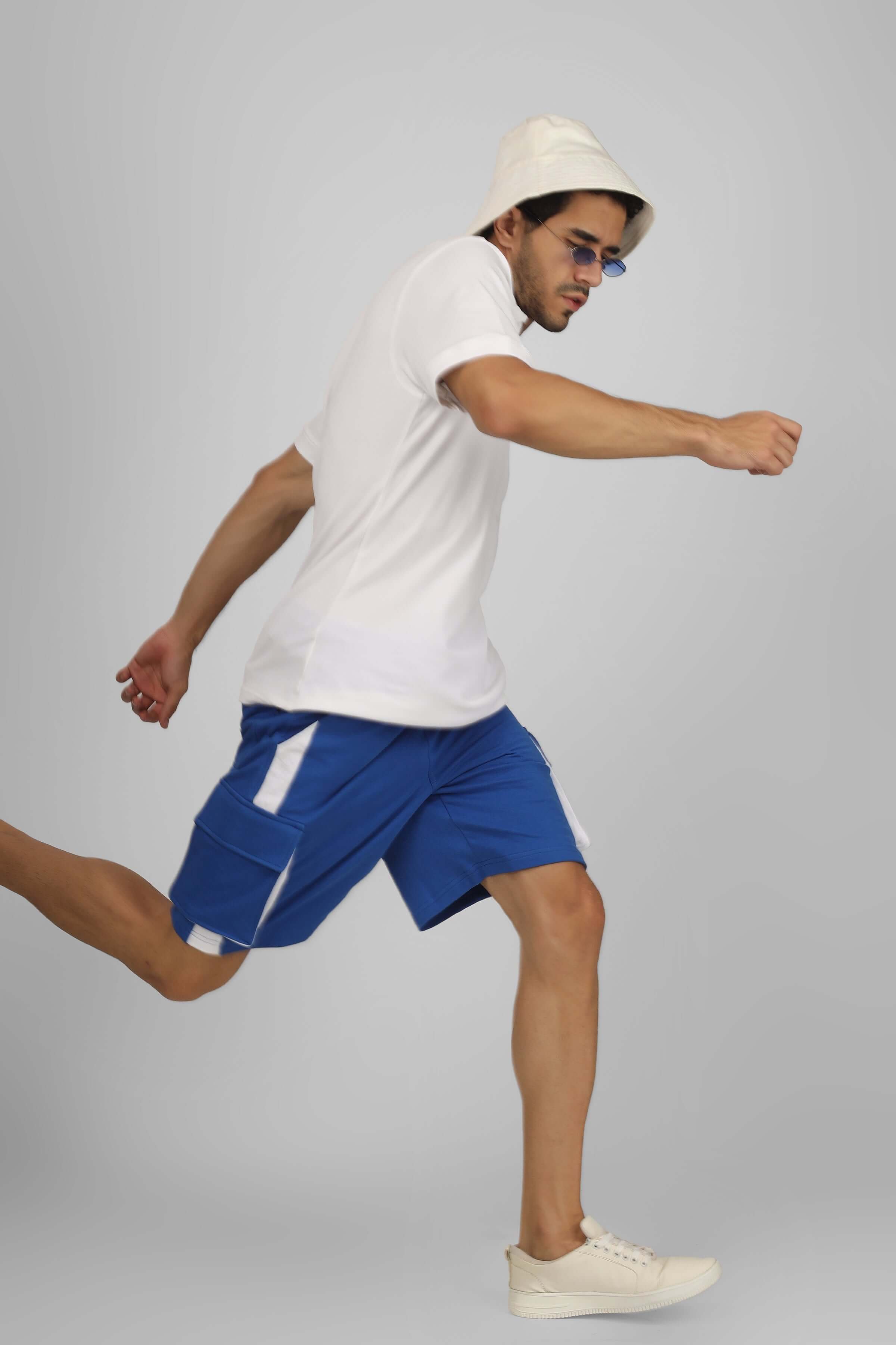 Man running in royal blue and white CONTRA cargo shorts, wearing a white t-shirt and hat, showcasing sporty and casual style.
