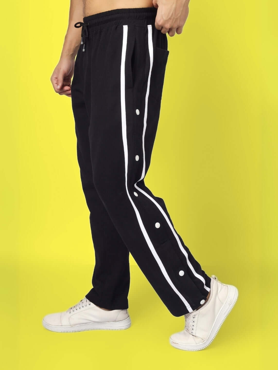 Side view of black contrast panel joggers with white stripes, modeled with white sneakers on a yellow background.