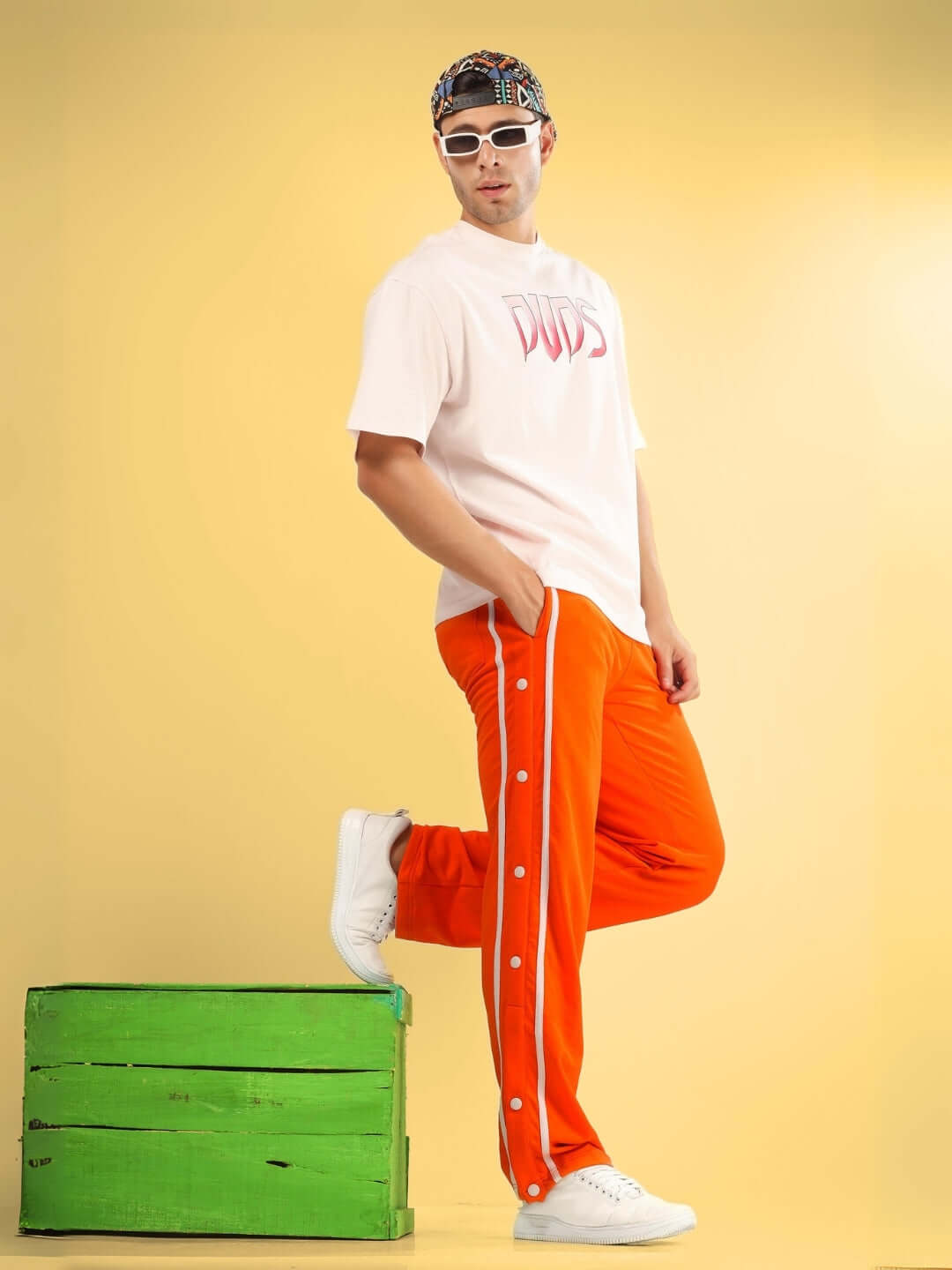 Model wearing orange contrast panel jogger pants with white sneakers, standing next to a green crate in a vibrant, stylish setting.